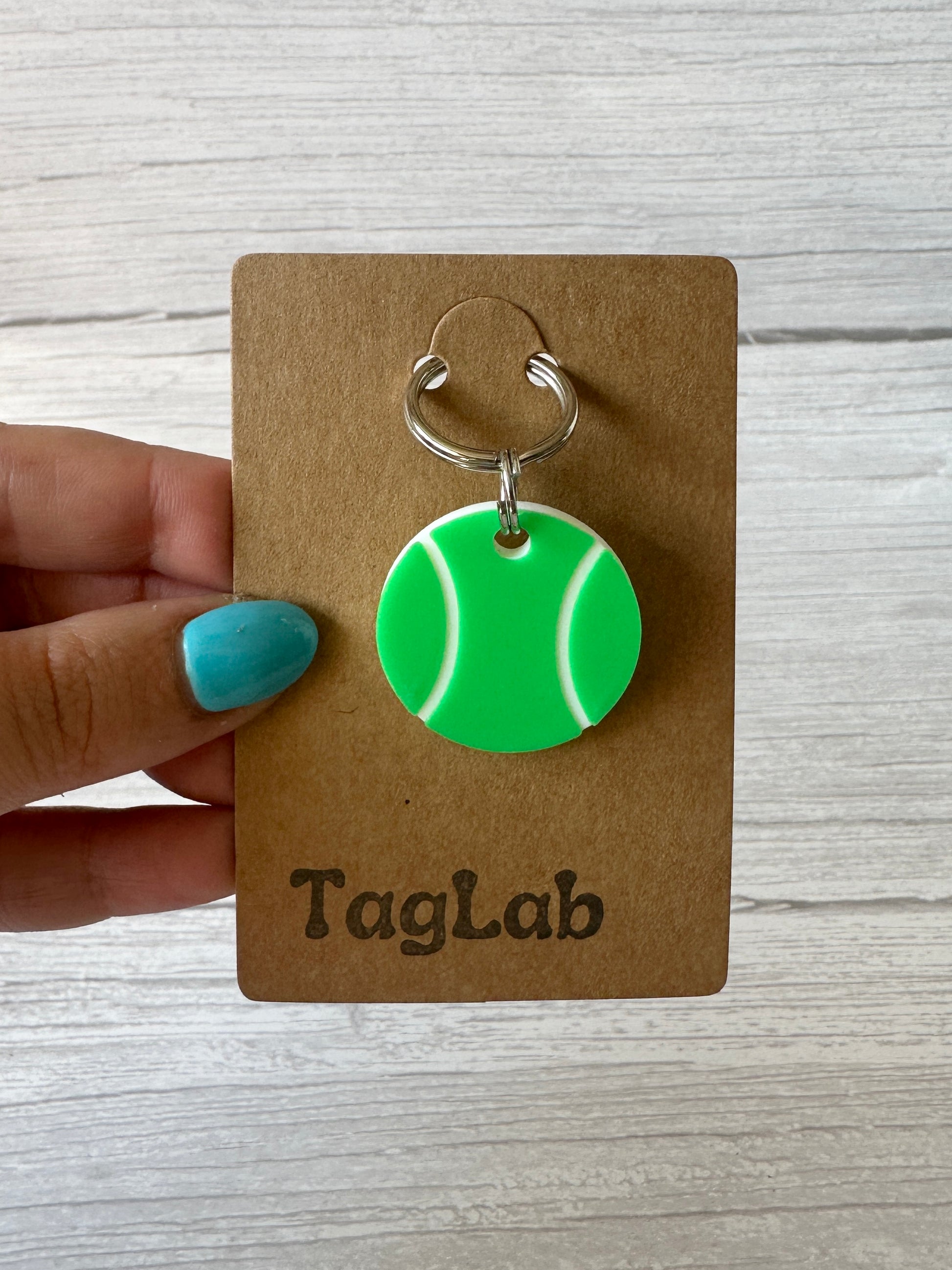 A hand with blue nail polish holds a brown "Crafts by Kate" card labeled "Ball is Life - Arnie & Mac’s Goodies," featuring a green, tennis ball-shaped pet tag. The background is a light wood surface.