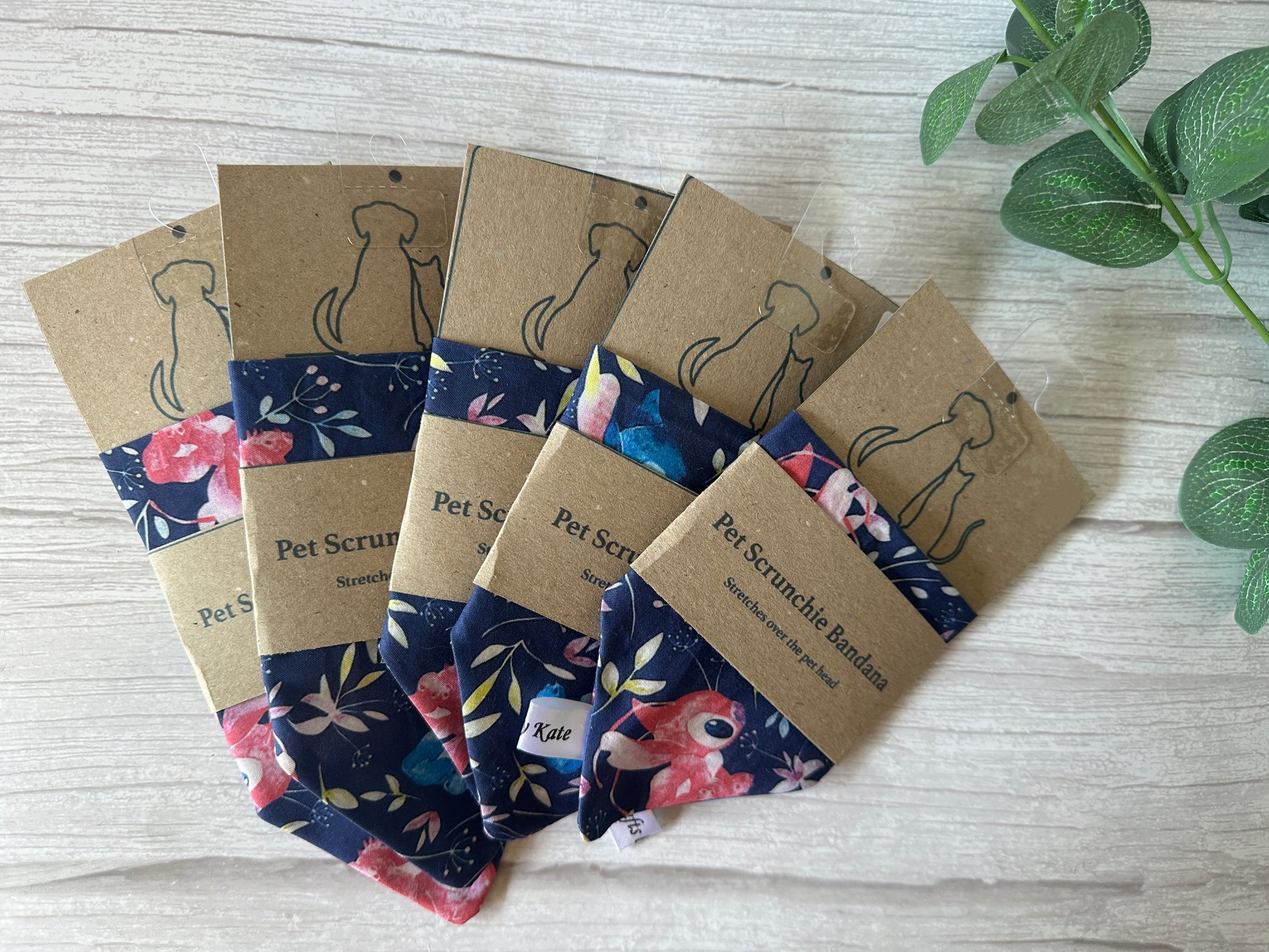 Five Cotton Pet Scrunchie Bandanas by Crafts by Kate, featuring floral designs from Stitch Obsession, are displayed in packaging on a light wood surface with a small green plant in the corner.