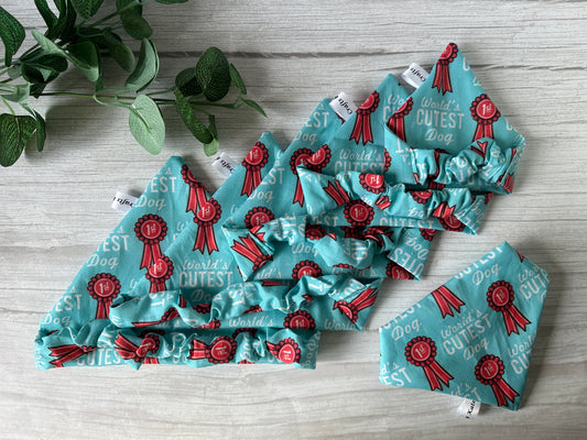 The Cotton Pet Scrunchie Bandana - World’s Cutest Dog by Crafts by Kate features red ribbon accents and "World’s Cutest Dog" text, shown in blue on a light wooden surface beside a green leafy plant.