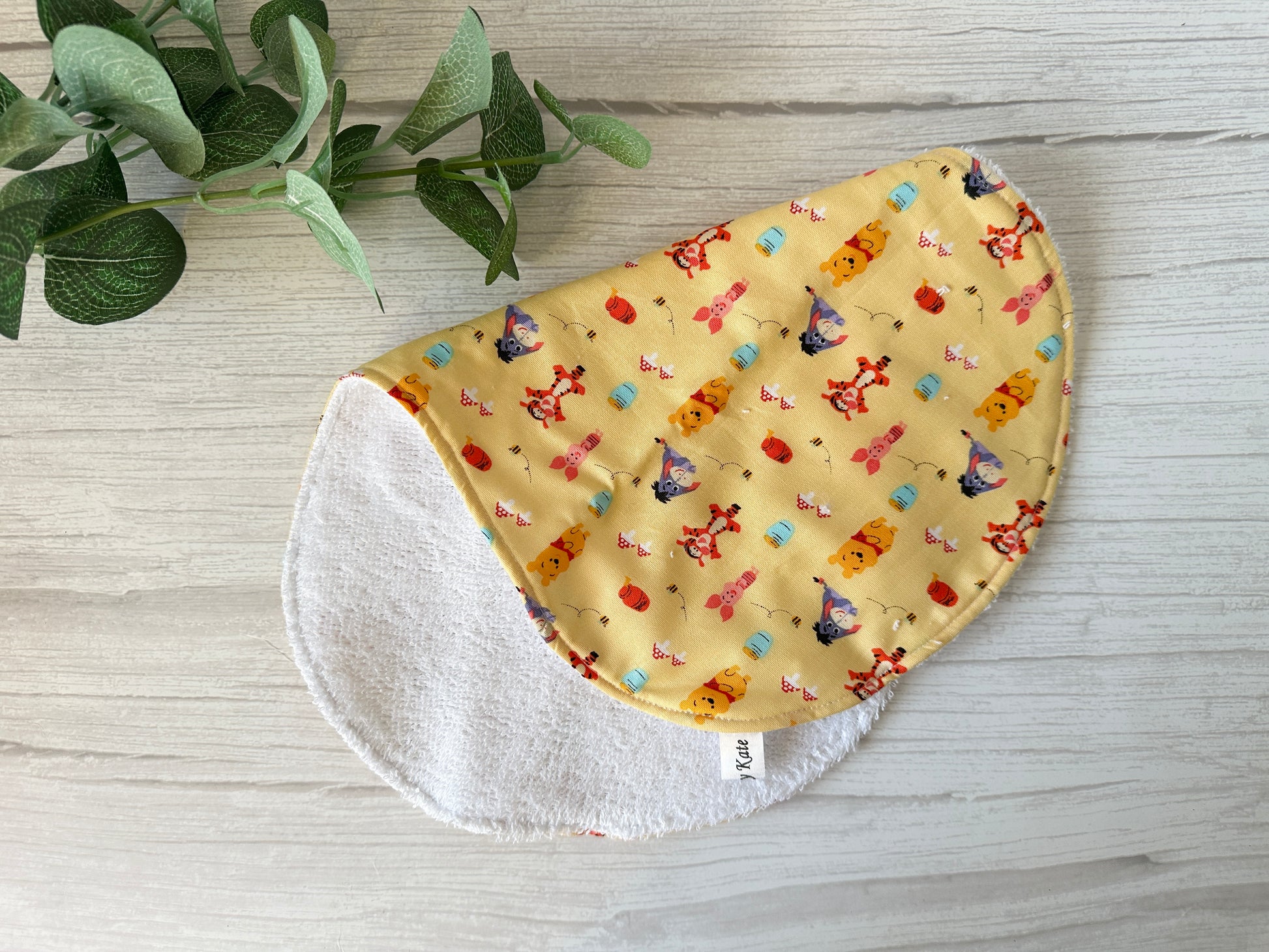 The Crafts by Kate Cotton & Terry Towelling Baby Gift Set features a round, two-sided ultra-soft cotton baby bib with colorful cartoon characters like Winnie the Pooh on yellow fabric, and a white terry cloth reverse. It sits on a light wooden surface next to a small green plant—perfect for any baby gift set.