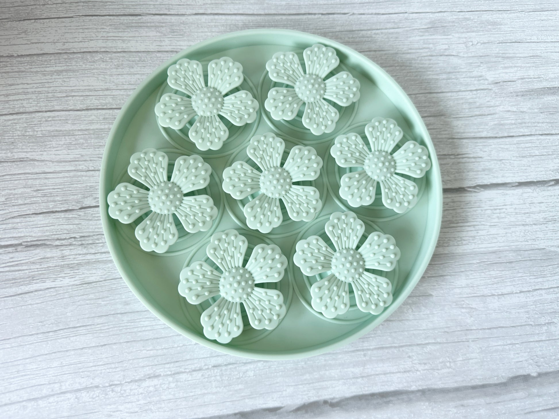 The Crafts by Kate Slow Feeder Pet Mat is a round, mint-colored tray featuring suction cups and securely holding six textured, mint flower shapes with detailed petals on a light wood background.