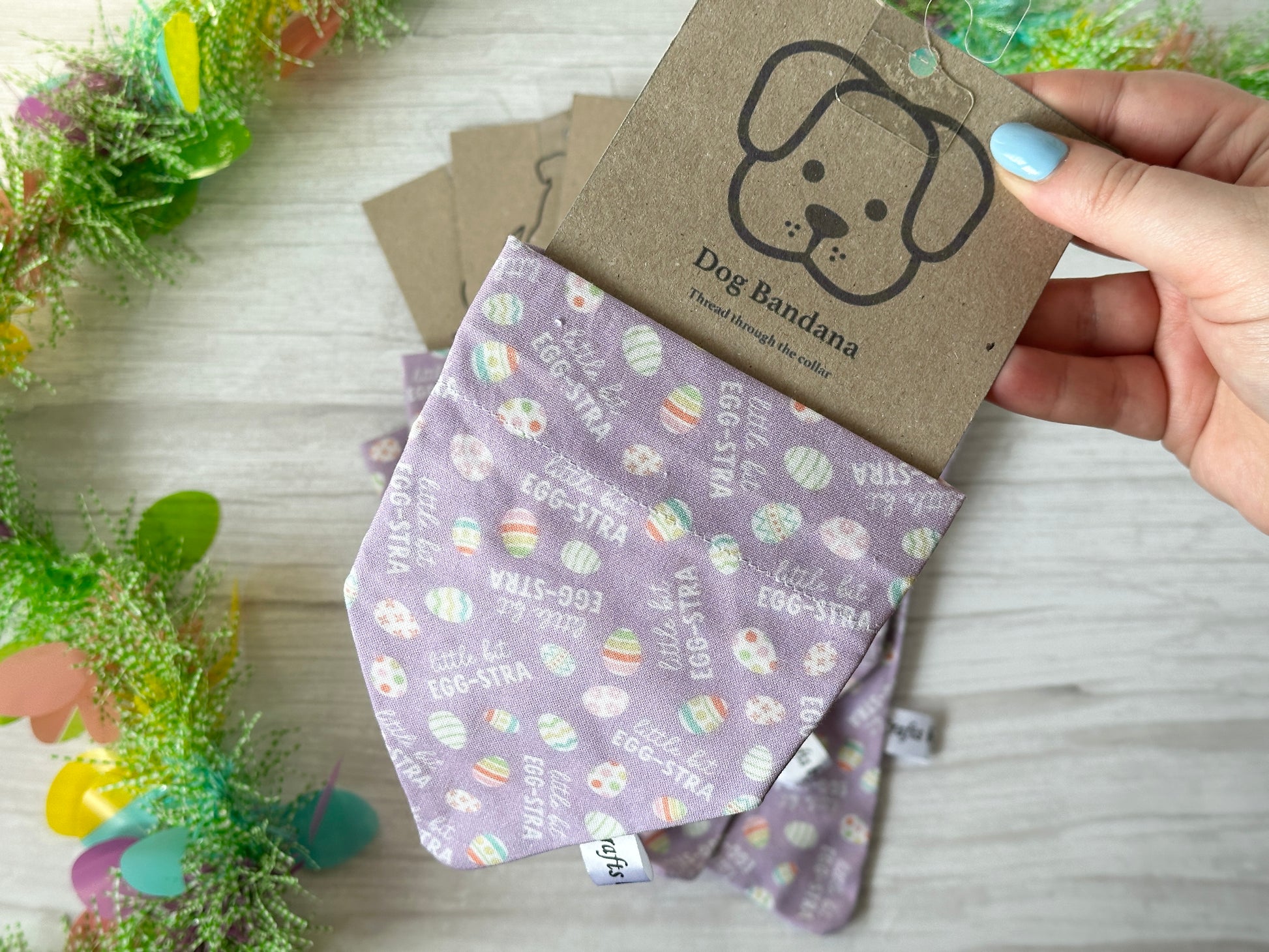 A hand holds a Crafts by Kate's "Little bit Egg-stra" Cotton Pet Bandana in purple, decorated with Easter eggs and phrases like "EGG-STRA" and "STAY EGG-STRA." It features a collar attachment for ease and is attached to a tag with a cute dog illustration and floral decorations.