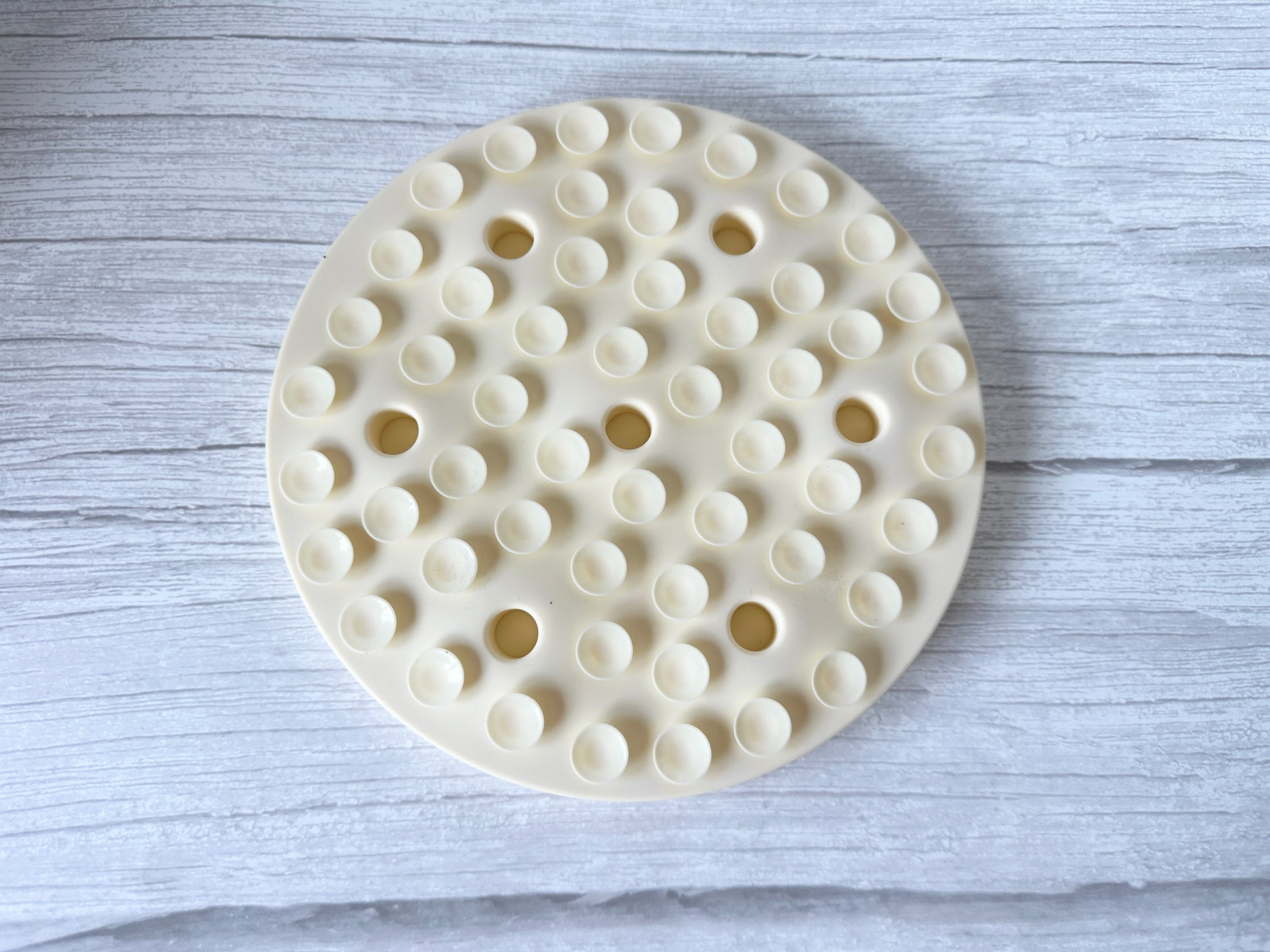The Crafts by Kate Slow Feeder Pet Mat is a round, beige plastic pegboard-like object with holes and bumps. It rests on a light gray wooden surface and features tiny suction cups underneath, making it a washable food mat that enhances pet digestion.