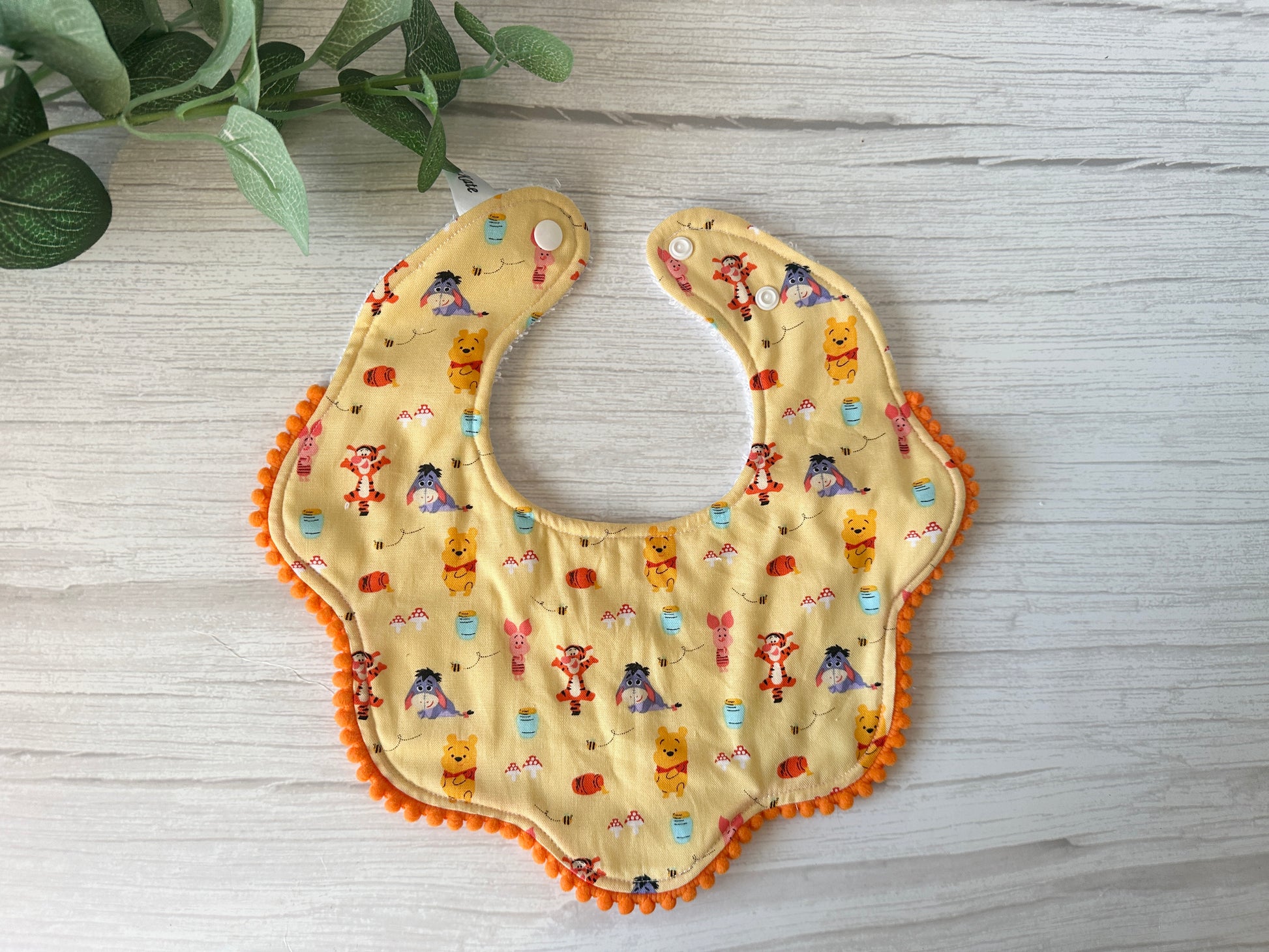 The Crafts by Kate Cotton & Terry Towelling Baby Gift Set features a Winnie the Pooh & Friends themed bib with playful animal cartoons on a cheerful yellow background, adorned with orange pom-pom trim, and set against a white wood surface accented with green leaves.