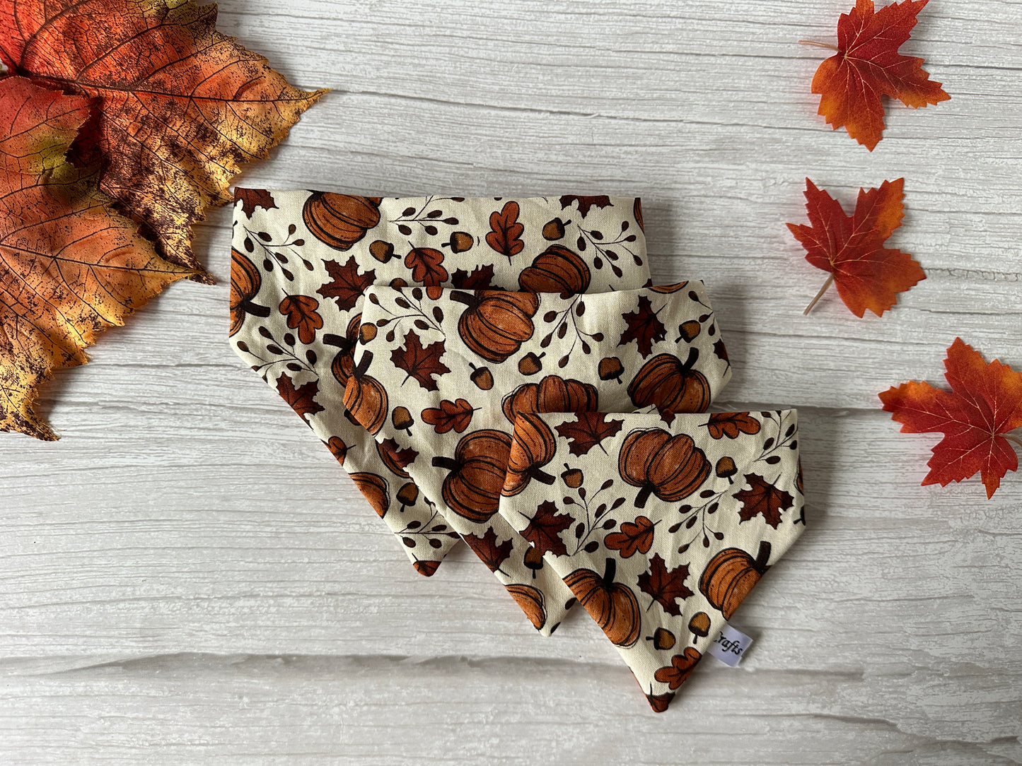 The Cotton Pet Scrunchie Bandana - Cottage Core Pumpkins by Crafts by Kate is gracefully arranged on a wooden surface, embodying an autumnal cottage core aesthetic. Surrounding the bandana are scattered red and orange maple leaves, accentuating the seasonal decor.