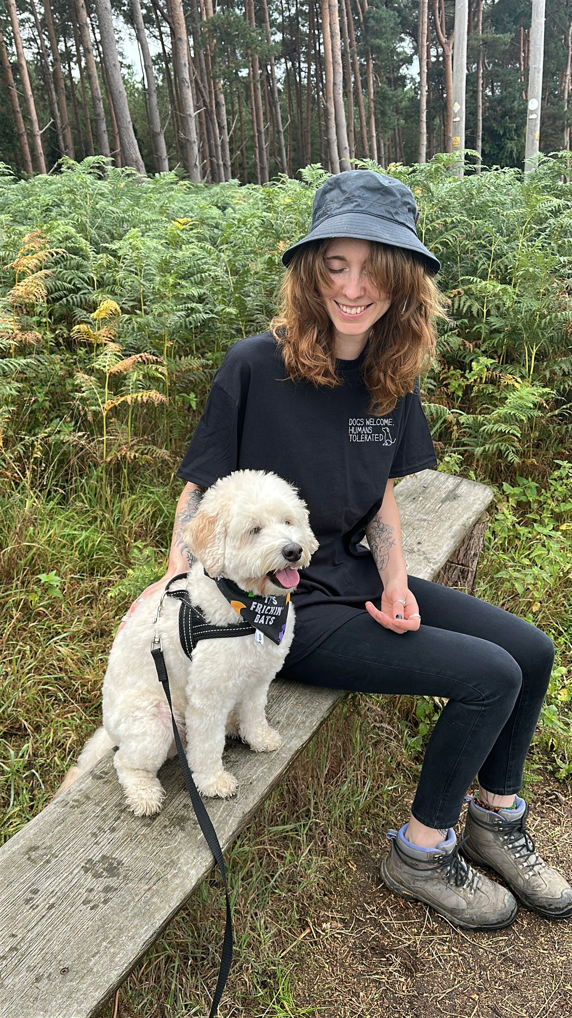 Wearing the "Dogs welcome" t-shirt by Crafts by Kate and a bucket hat, a smiling woman with wavy hair sits in a forest with her fluffy white dog, surrounded by green ferns and tall trees.