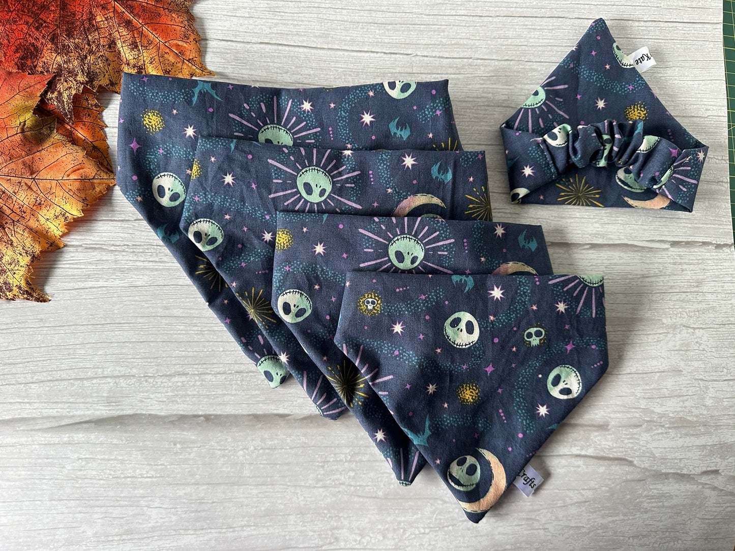 A set of four dark-themed fabric bandanas, featuring celestial and spooky designs like suns, moons, and skulls, are neatly arranged on a light wooden surface. Nearby, a Cotton Pet Scrunchie Bandana - Jack Skeleton by Crafts by Kate and a large autumn leaf add to the display.