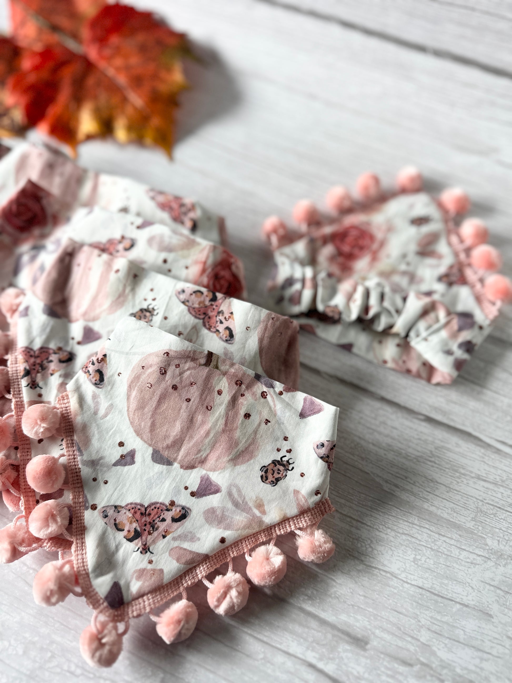 A set of decorative, pom-pom trimmed fabric bunting pieces featuring pastel-colored designs of pumpkins and butterflies, arranged on a light wooden surface. A red and orange autumn leaf is partially visible in the background, adding a touch of seasonal charm to the Cotton Pet Scrunchie Bandana - Roses & Pumpkins from Crafts by Kate.