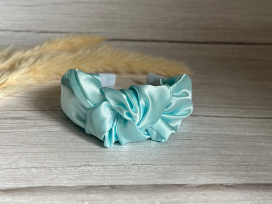 The Silk Knotted Headband - Aqua Blue from Crafts by Kate sits elegantly on a wooden surface, its handcrafted silk design featuring a sophisticated knot. The background showcases beige, feathery decorative elements, exuding an air of luxury.