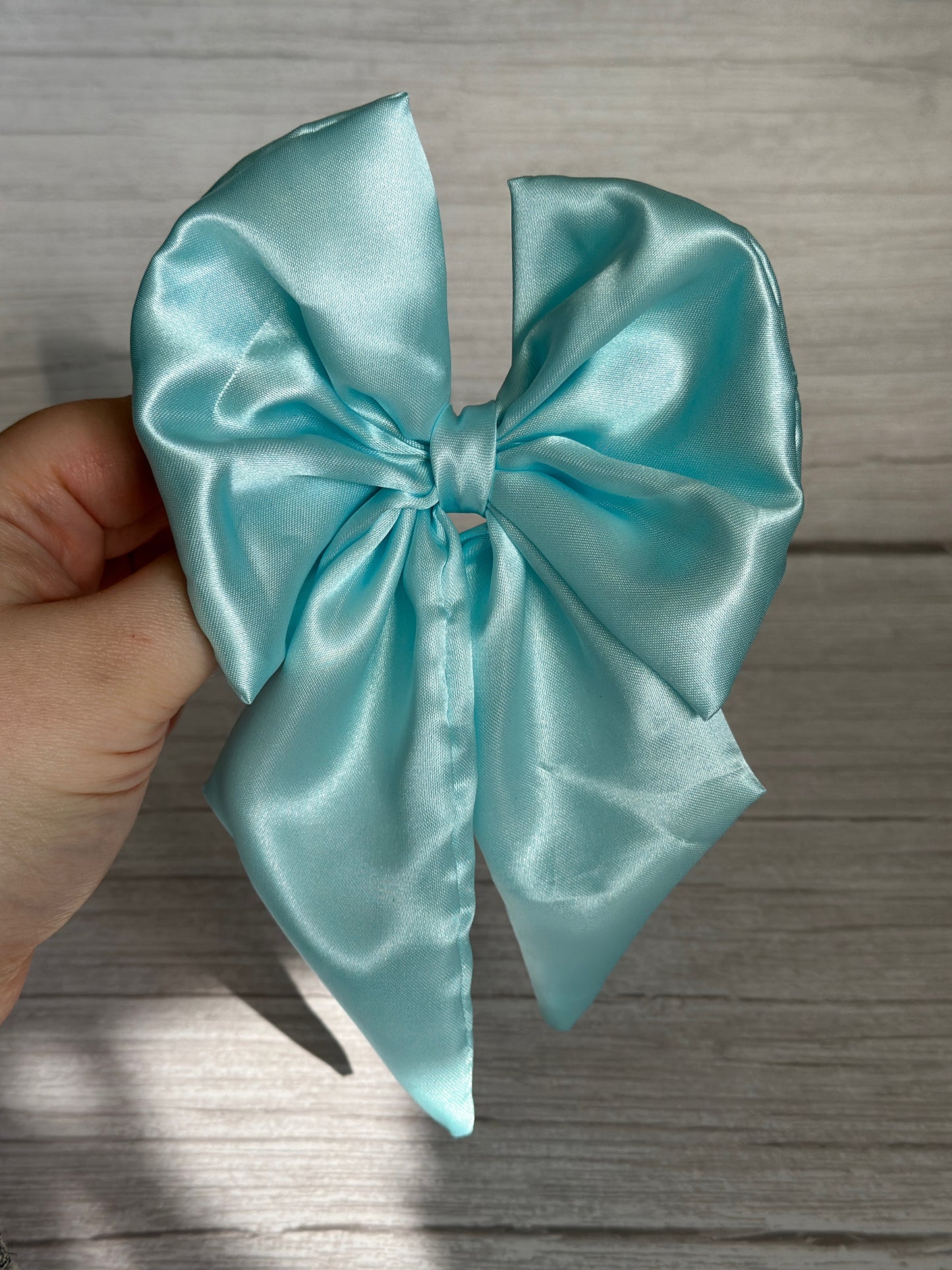 A hand delicately holding a large, shiny Silk Sailor Bow Clip in Aqua Blue from Crafts by Kate against a wooden background. This luxurious clip is expertly handcrafted and neatly tied with long, elegant tails.