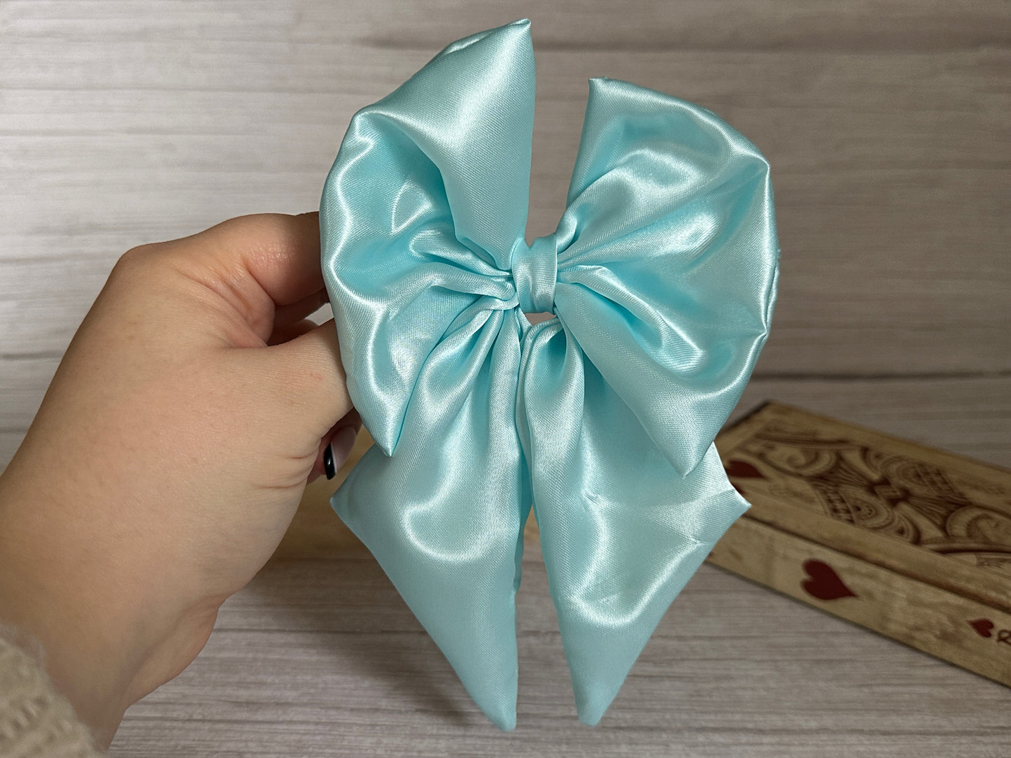 A hand holds the elegant Silk Sailor Bow Clip - Aqua Blue by Crafts by Kate against a wooden background with decorative engravings. The luxurious handcrafted bow boasts two long tails and a tied center.