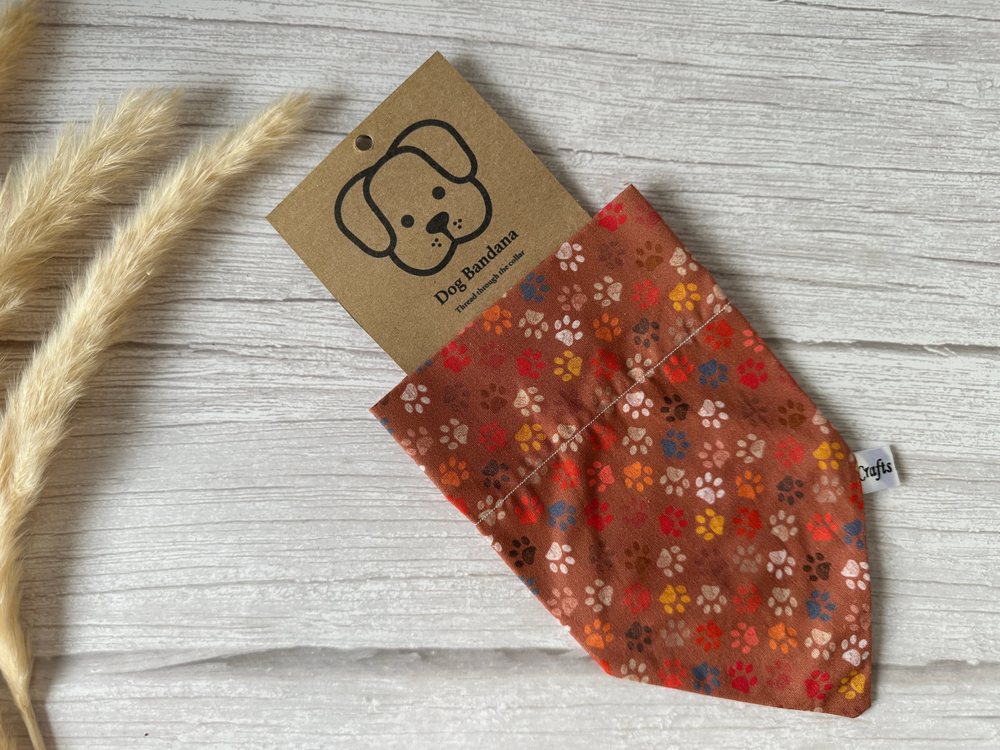 An Autumn Paws bandana with a multicolored paw print pattern, labeled "Cotton Pet Bandana with collar attachment - Crafts by Kate," is partially pulled out from its packaging. The packaging features a simple drawing of a dog's face and the text "Dog Bandana." Dried wheat stalks are placed beside the bandana on a wooden surface.