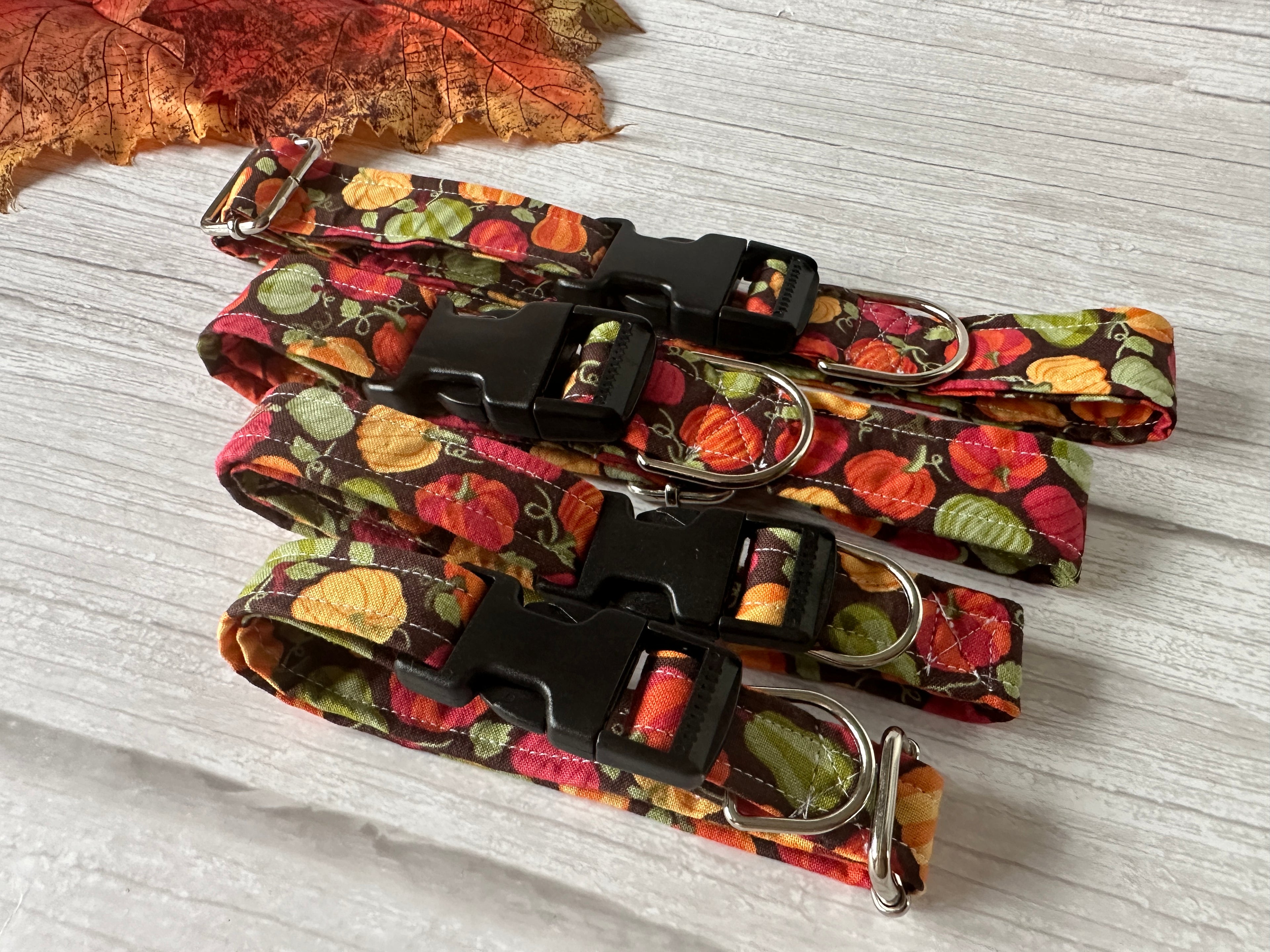 Three "Pumpkin Lover" Cotton Pet Adjustable Collars from Crafts by Kate are laid out on a light wooden surface. The autumn-themed collars feature colorful pumpkins and leaves, and are adorned with silver metal hardware. They are aligned in a row, with a decorative autumn leaf partially visible in the upper left corner.