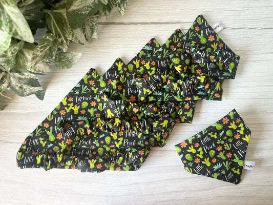 Five handcrafted black Cotton Pet Scrunchie Bandanas by Crafts by Kate, featuring colorful cactus and "Little Prick" patterns, are arranged on light wood with green plants—perfect accessories for stylish pets.