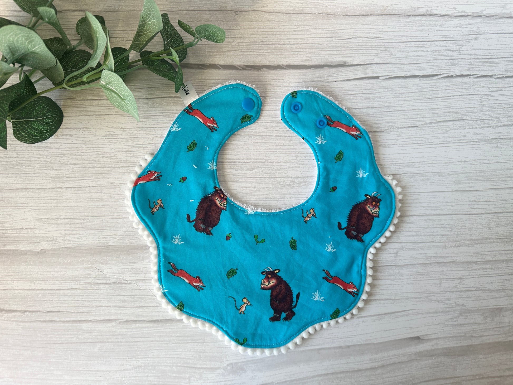 The Gruffalo in Blue baby gift set by Crafts by Kate includes a blue bib with mythical creatures, foxes, and leaves. Made from ultra-soft cotton with scalloped edges and dual snap buttons, it's ideal for a handcrafted collection. Features a small green plant in the upper left corner on light wood.