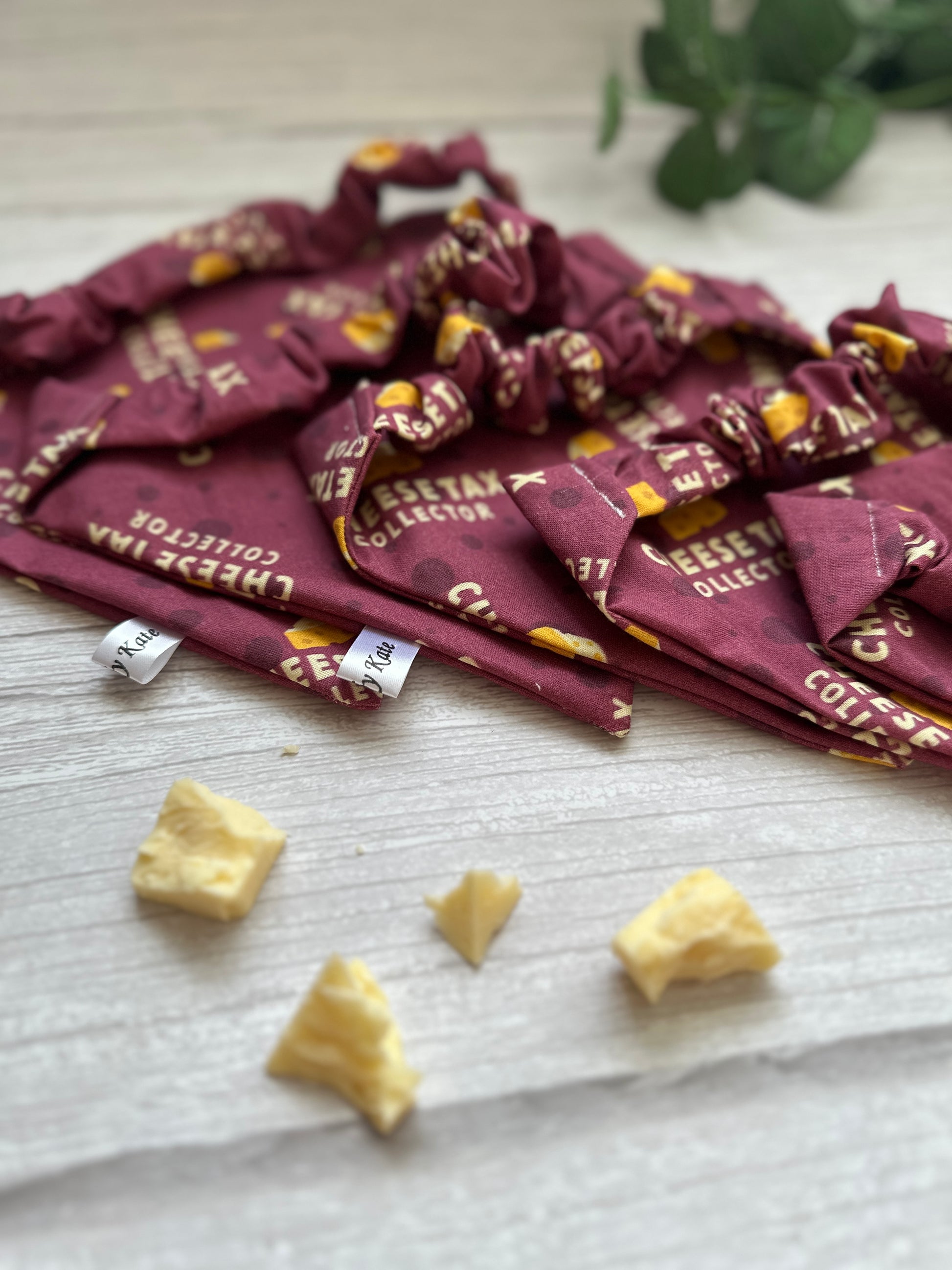 Crafts by Kate's Cotton Pet Scrunchie Bandana - Cheesetax Collector bags, containing small cheese pieces, are arranged on light wood. A few cheese bits and a hint of green plant are visible in the background, echoing the vibrant energy of a handcrafted bandana collection.