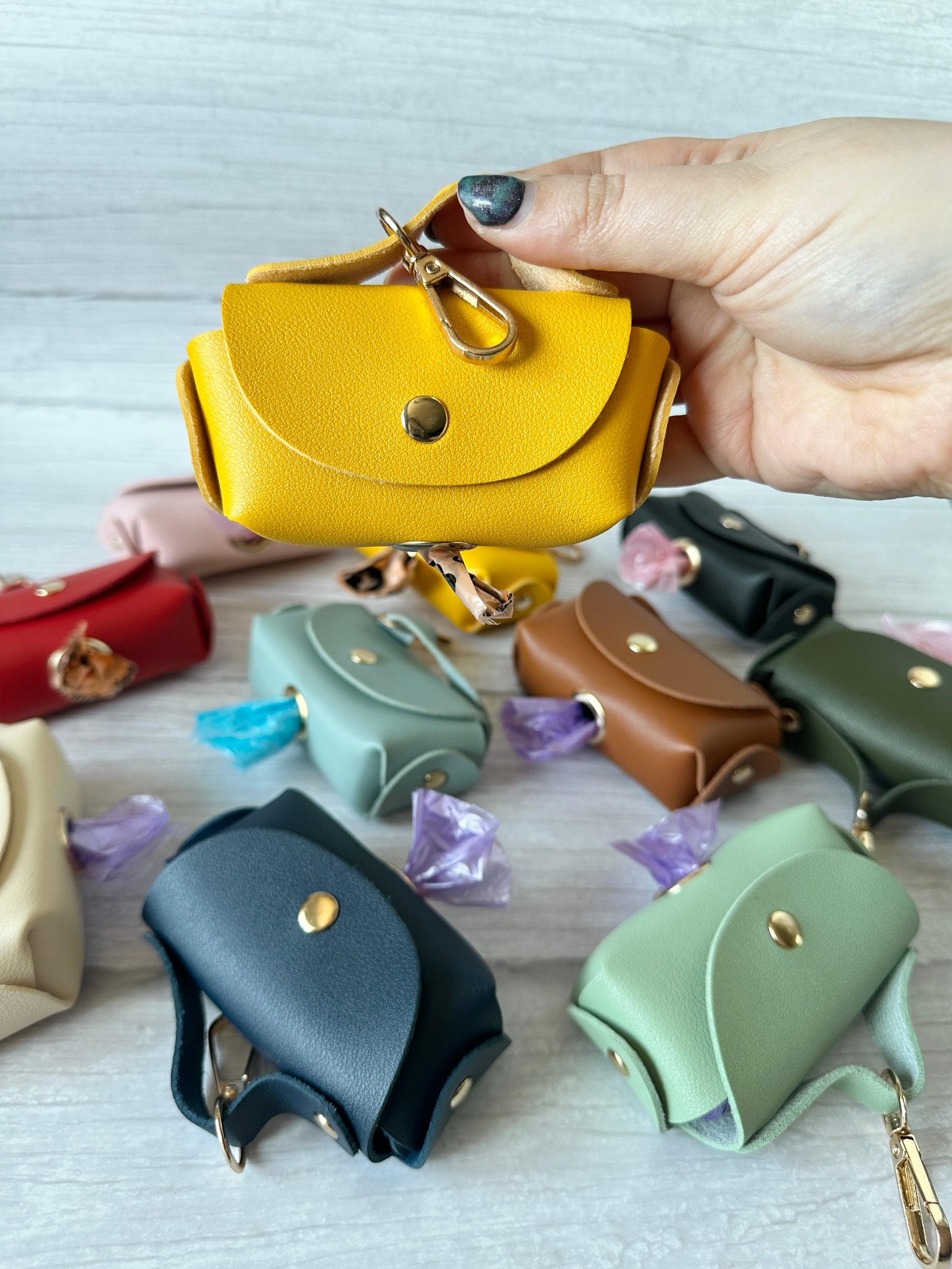 A hand displays a yellow leather "Pet Poo Bag Holder Handbag" by Crafts by Kate, surrounded by colorful faux leather holders in pink, blue, brown, and green on a light wood surface. Purple wrapping paper is visible between them.
