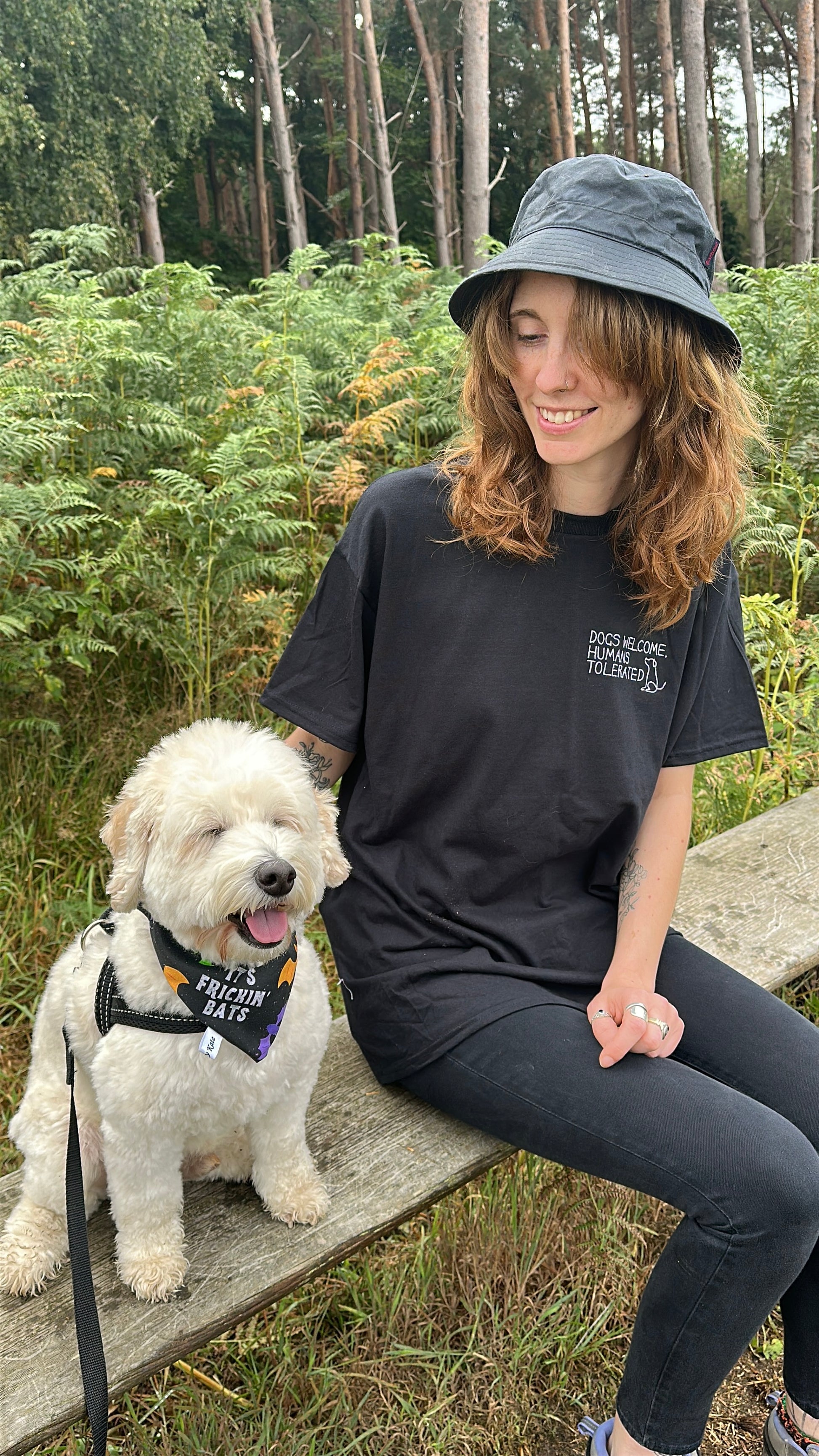 A woman with wavy brown hair, wearing a black Dogs welcome t-shirt by Crafts by Kate and a black hat, sits outdoors on a wooden bench beside a small white dog in a “FREAKIN’ BATS!” harness. Green foliage and trees fill the background.