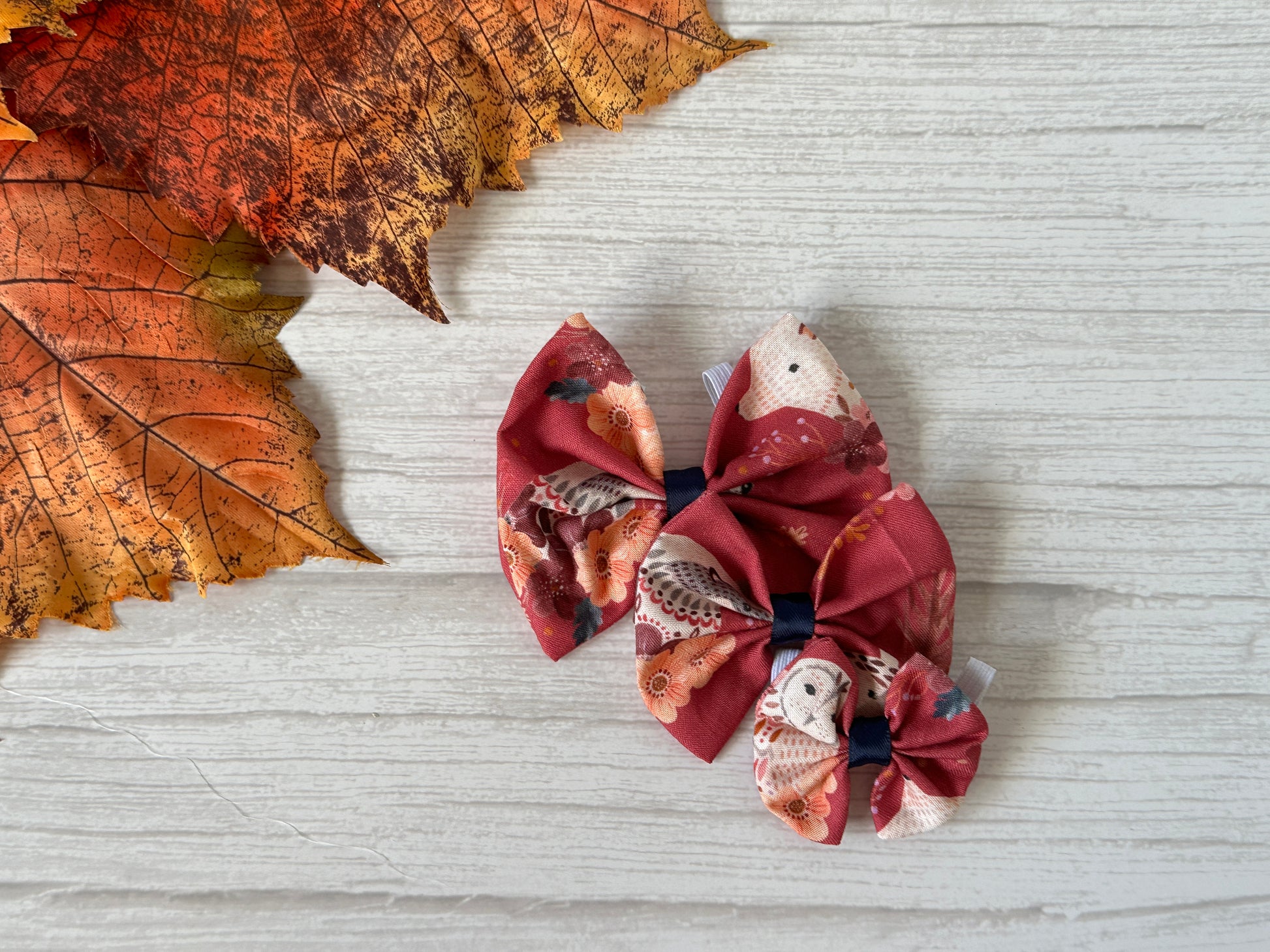 A collection of autumn-themed hair accessories from Crafts by Kate, featuring the Cotton Pet Bow with collar attachment in a woodland animals design, is displayed on a white wooden surface. Nearby, orange and yellow fall leaves accentuate the seasonal theme.