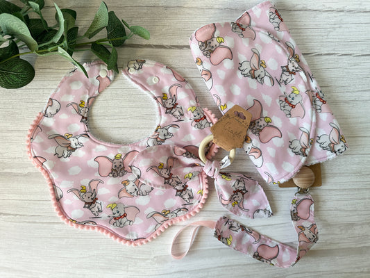 The Cotton & Terry Towelling Baby Gift Set - Dumbo in pink by Crafts by Kate includes a soft cotton bib, pacifier clip, and blanket with a cartoon elephant design on a pink background, complemented by a small potted plant on a light wooden surface.