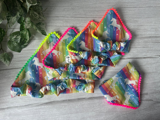 Four vibrant “Bitch I’m a Unicorn” Cotton Pet Scrunchie Bandanas by Crafts by Kate, featuring rainbow stripes and unicorn silhouettes, are arranged on a light wooden surface beside a green leafy plant.