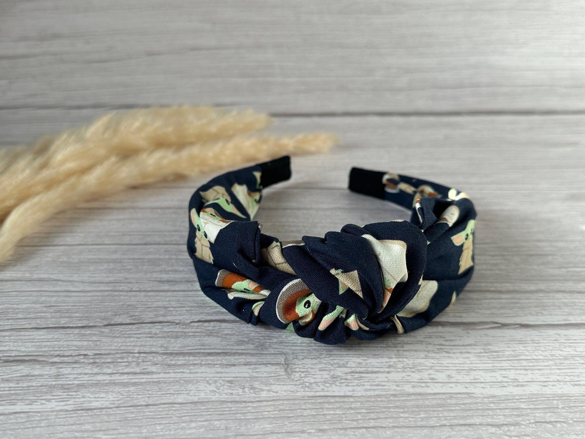 A navy blue, handcrafted luxury statement headband with a twisted knot at the center, featuring a colorful floral pattern. The Crafts by Kate Cotton Knotted Headband - Baby Yoda is placed on a light wood background, with some dried pampas grass to the left.