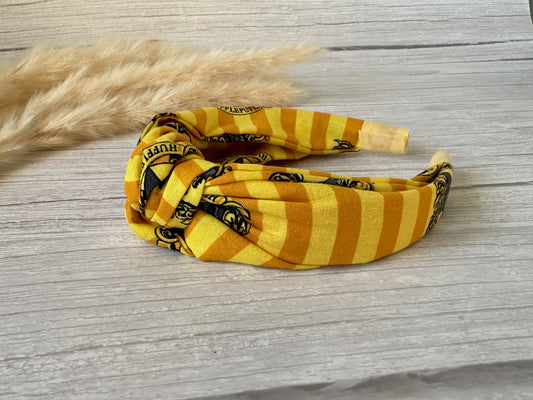 A luxurious Cotton Knotted Headband from Crafts by Kate, striped in yellow and orange with black Hufflepuff text and illustrations, rests on a light wooden surface. Fluffy beige pampas grass decorates the background, adding a cozy elegance to the scene.