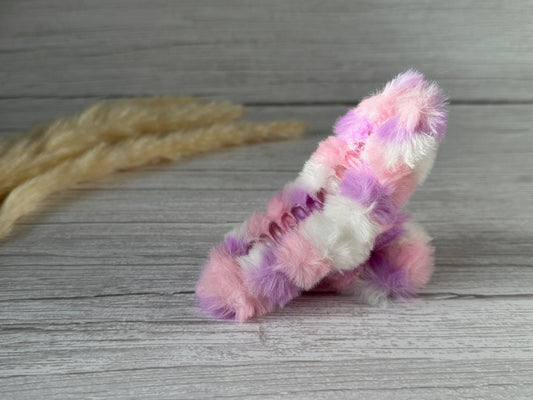 A plush, multicolored toy rocket made of luxury plush fluff fabric lies on a wooden surface. The toy features a mix of pastel colors including pink, purple, and white. In the background, light beige dried plant stems are slightly blurred. Nearby, Crafts by Kate's Fluffy Hair Claw Clip - Barbie Fantasy adds an extra touch of elegance.