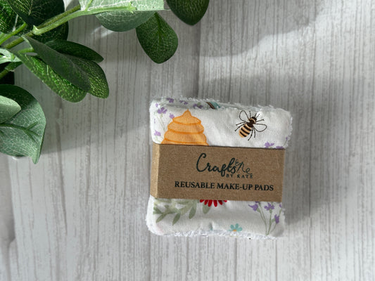 A set of "Cotton Reusable Make-Up Remover Pads - Bee Garden" wrapped in a Kraft paper band with the branding "Crafts by Kate." These handcrafted cotton pads feature colorful prints of bees, flowers, and honeycombs. They are placed on a light wood surface with a small leafy plant nearby.