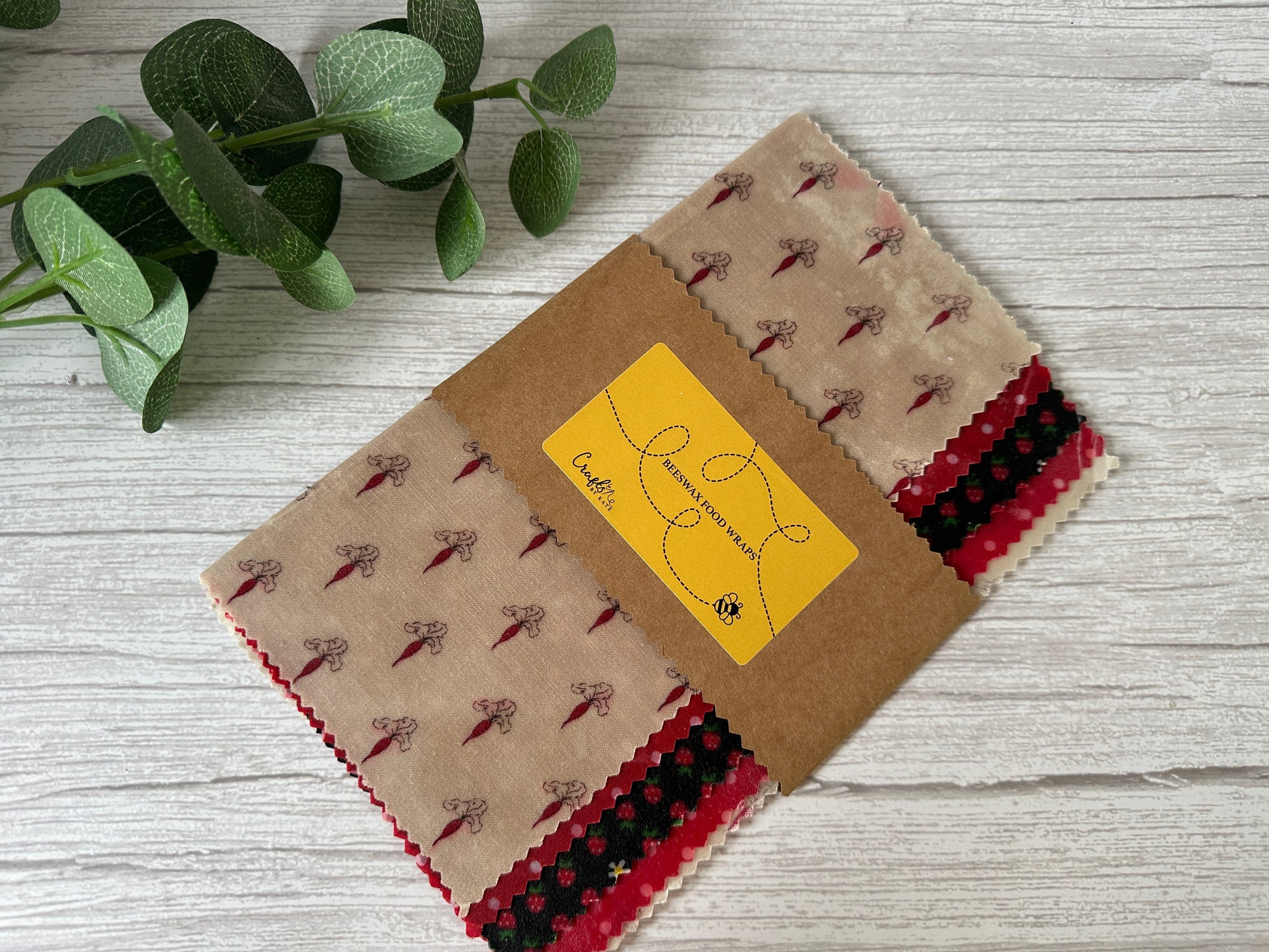 A set of Crafts by Kate Beeswax Food Wraps, adorned with pink floral and checkered patterns, is neatly folded and secured with a brown kraft paper sleeve on a light wooden surface. These organic wraps are designed to help keep your food fresh. A small leafy plant can be seen in the top left corner of the image.