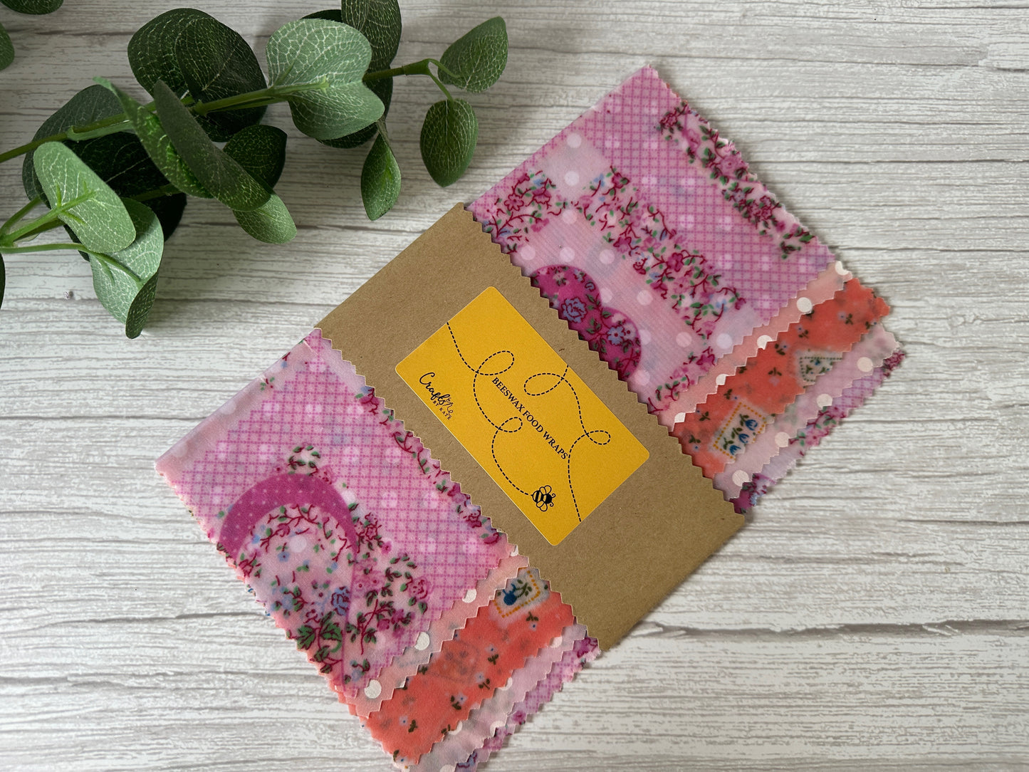 A beige organic beeswax food wrap from Crafts by Kate, featuring a red radish pattern and a smaller red, black, and green striped section, is neatly folded in brown paper packaging labeled with a yellow tag. Renowned for keeping food fresh, it is set against a green leafy plant on a light wooden surface.