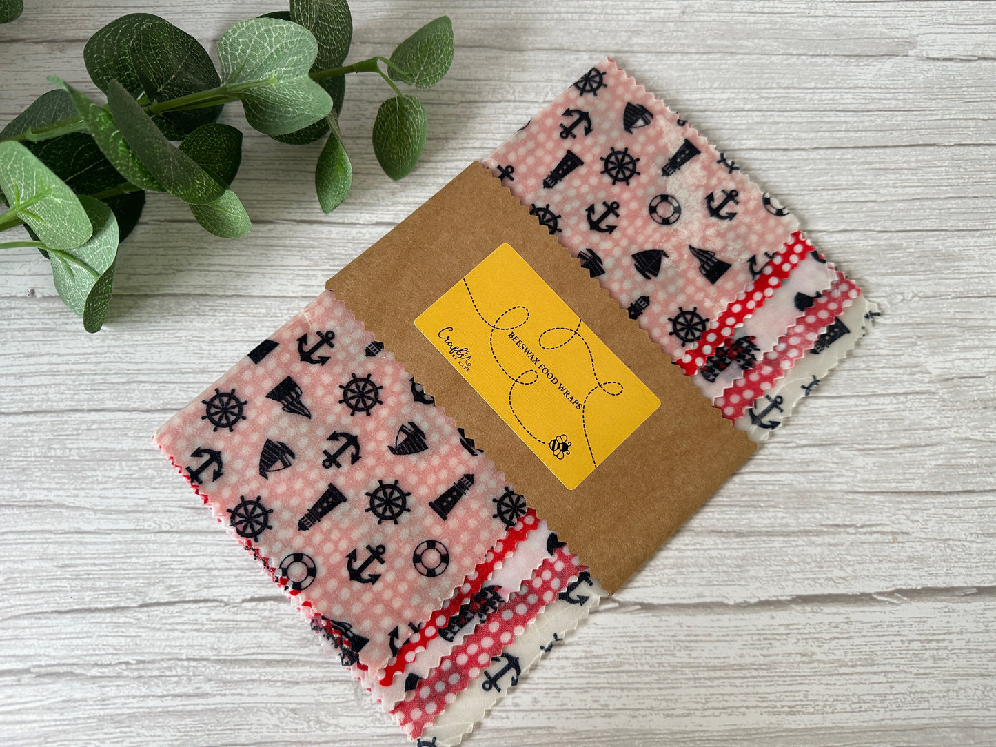 A bundle of colorful fabric swatches is held together with a brown paper band. The top visible swatch features a red and pink rose pattern alongside a black and white houndstooth design. Adjacent to the fabrics, a Beeswax Food Wrap by Crafts by Kate rests on a light wooden surface next to a leafy green sprig.