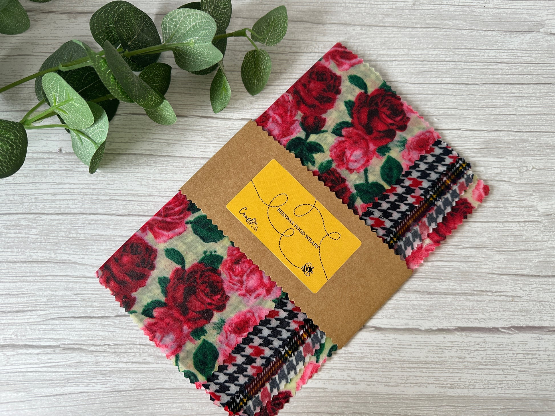 A bundle of colorful fabric swatches is held together with a brown paper band. The top visible swatch features a red and pink rose pattern alongside a black and white houndstooth design. Adjacent to the fabrics, a Beeswax Food Wrap by Crafts by Kate rests on a light wooden surface next to a leafy green sprig.