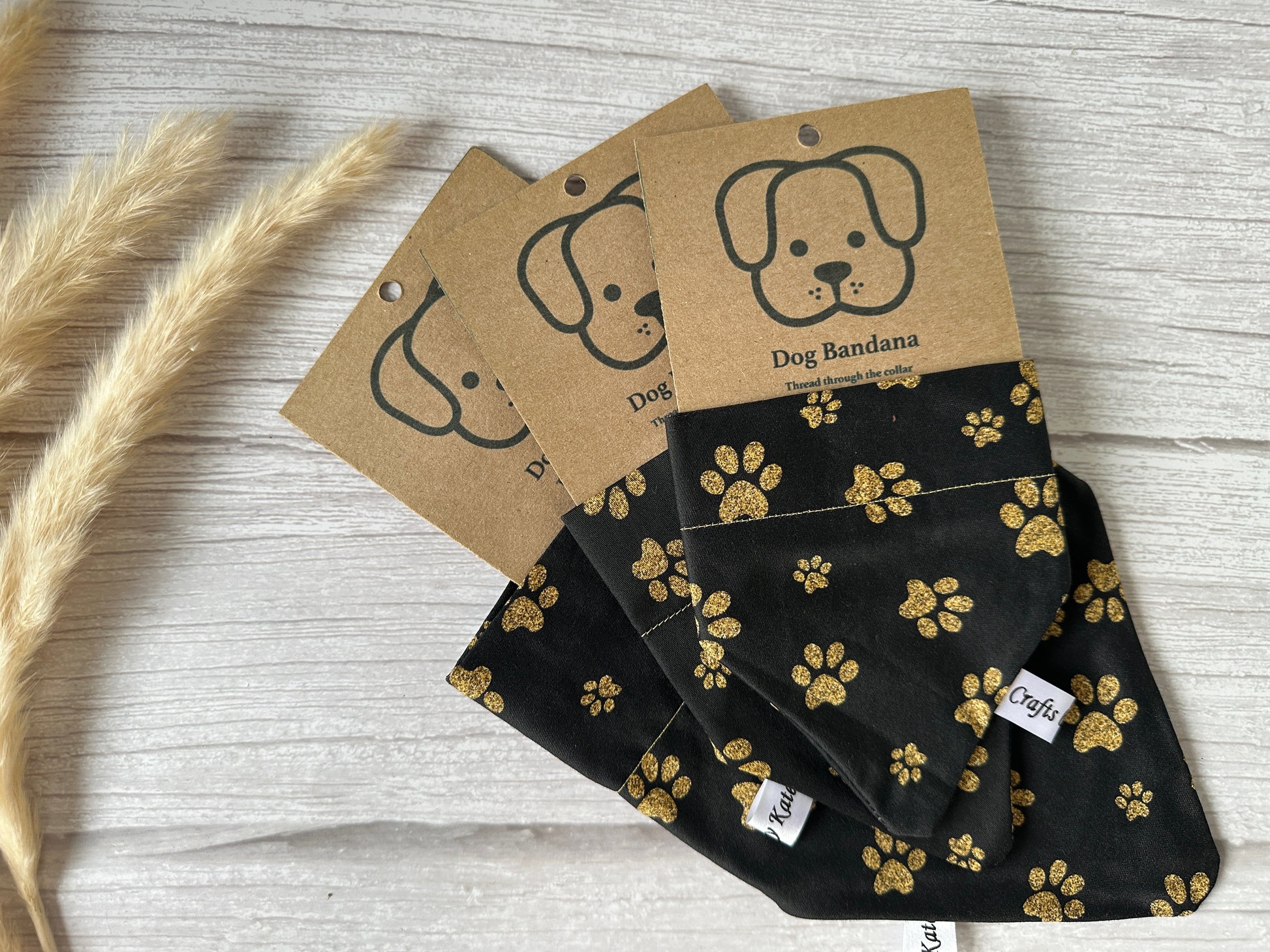 Three Cotton Pet Bandanas, each featuring the Black & Gold Paws design from Crafts by Kate, are laid on a light wooden surface. Each bandana is designed to easily slide through a pet collar and is attached to a brown cardboard tag with an illustration of a dog and the text "Dog Bandana." Wheat stalks are visible on the side.