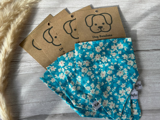 Several handcrafted Cotton Pet Bandanas with collar attachments from Crafts by Kate are displayed, each featuring a label with a simple dog illustration and the text "Dog Bandana." Made from Blue Daisies fabric, the bandanas are arranged in a fan shape on a white wooden surface with dried pampas grass beside them.