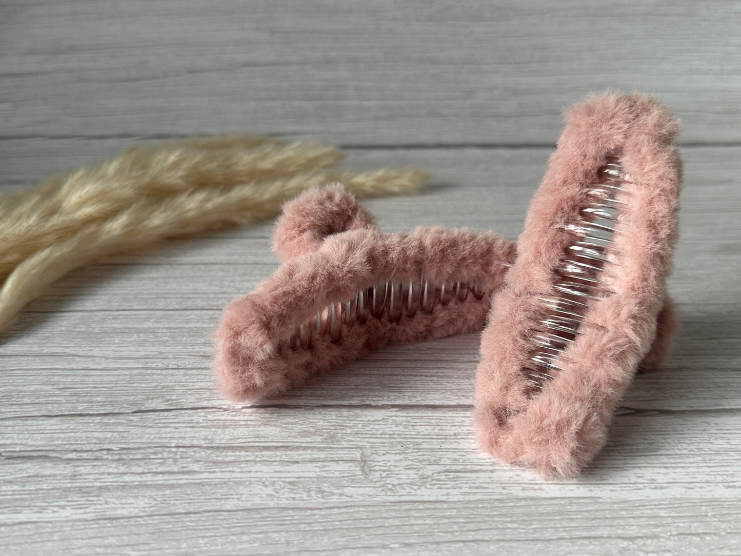 Two Crafts by Kate "Fluffy Hair Claw Clips - Blush Pink" with silver teeth are positioned on a light wooden surface. In the background, beige, dried decorative plants add a natural touch to the composition. The scene is softly lit, enhancing the luxurious plush fluff fabric of these handcrafted accessories.