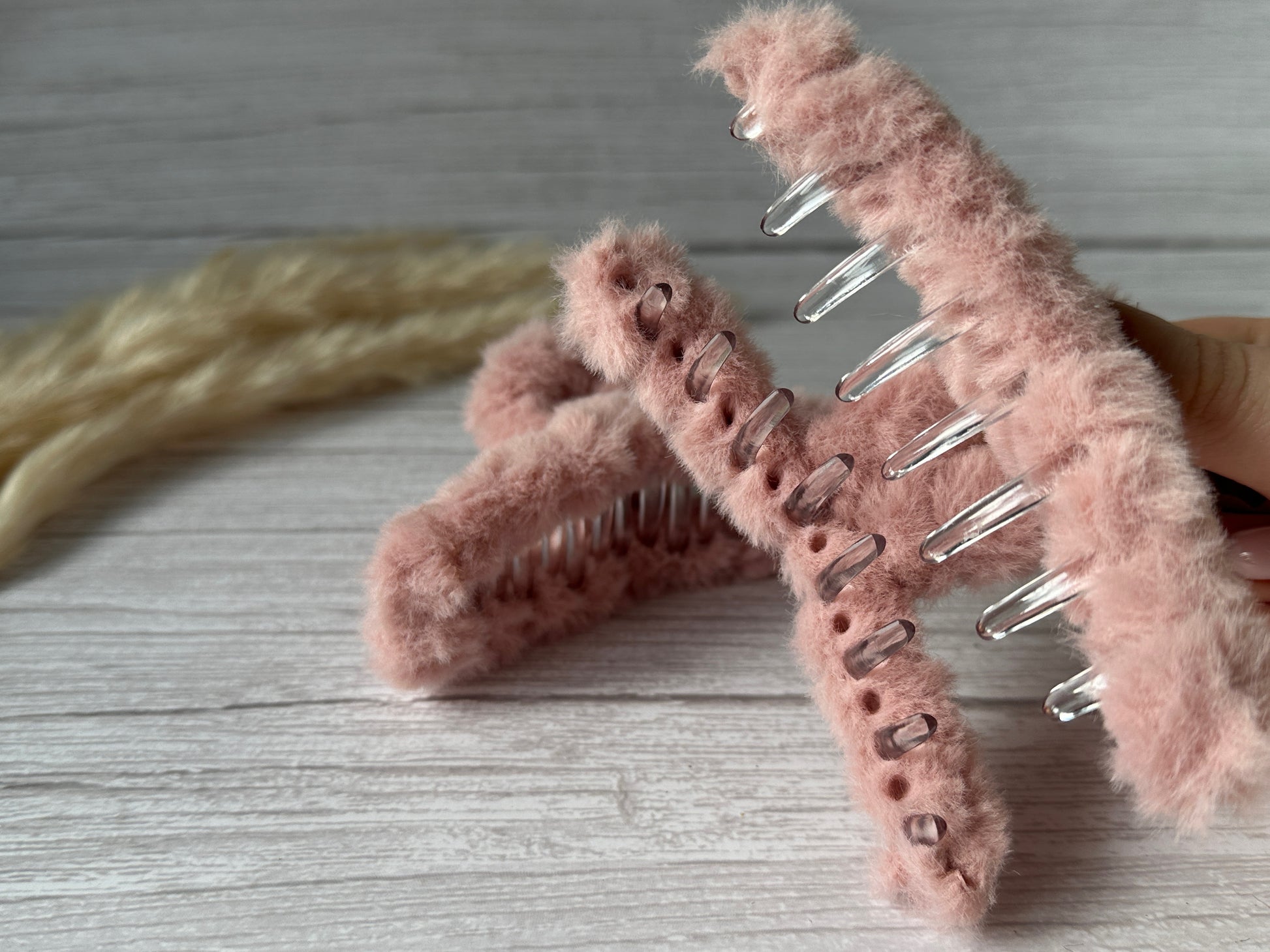 Close-up of a hand holding the Crafts by Kate Fluffy Hair Claw Clip in Blush Pink, made from luxury plush fluff fabric, against a light wooden background. Another handcrafted fuzzy clip and a braid of blonde hair are also visible. The clips feature a metal base covered with soft, textured fabric on the outer surface.