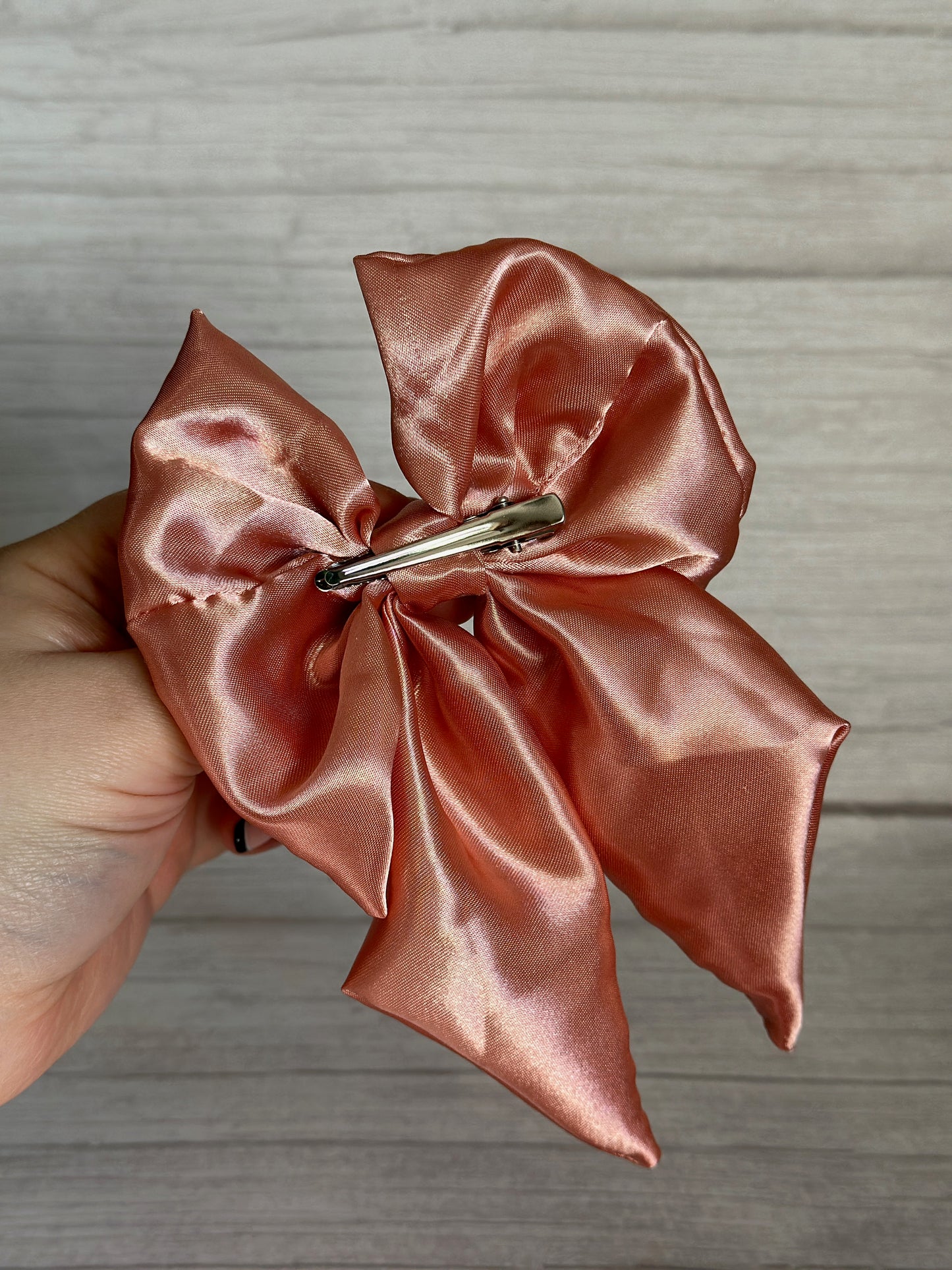 A hand cradles the Crafts by Kate Silk Sailor Bow Clip in blush pink satin, its metal clip gleaming. The bow unfurls elegantly, highlighting its texture and sheen against a wooden backdrop.