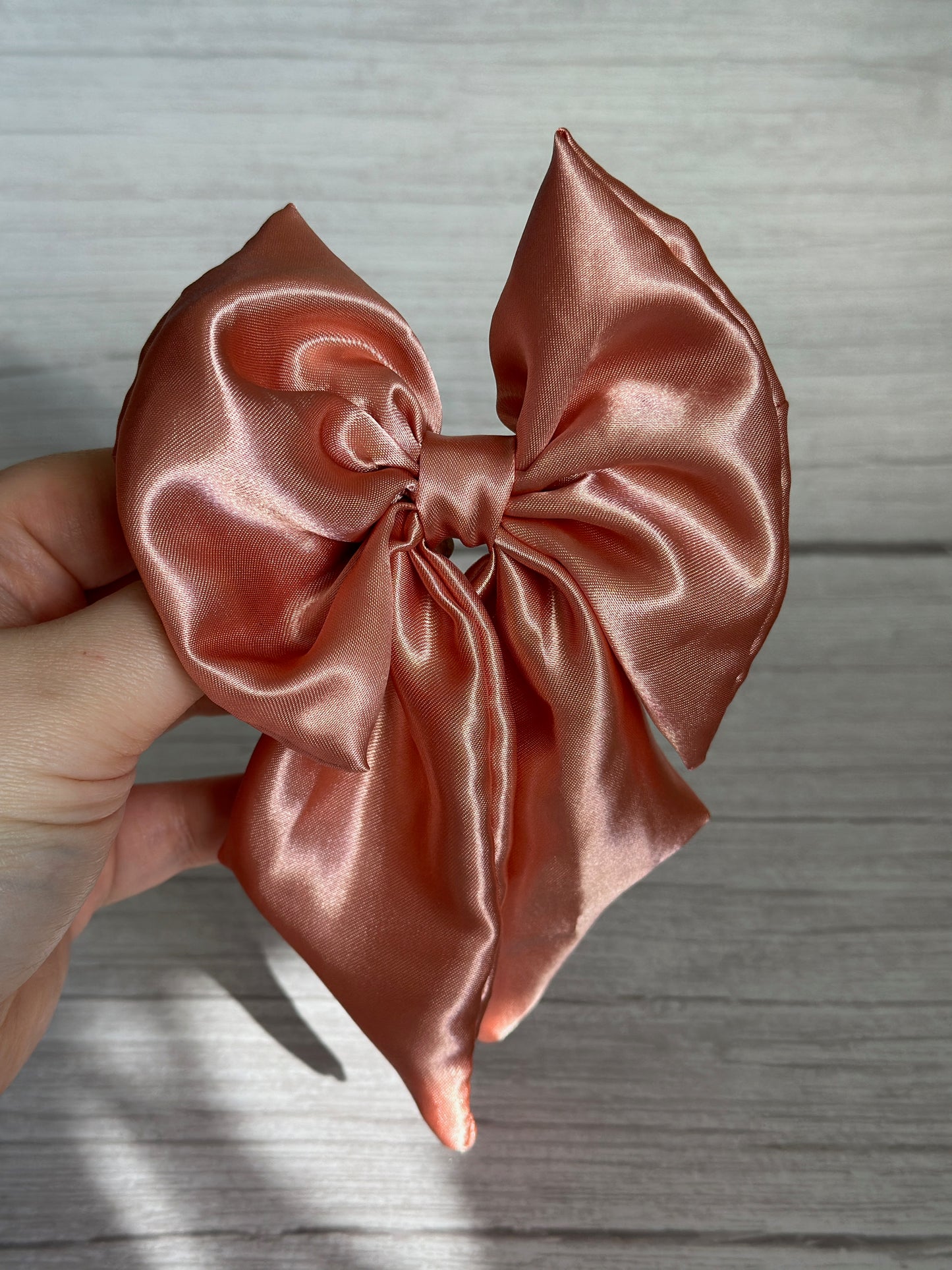 A hand grasps the elegant Silk Sailor Bow Clip in a beautiful blush pink shade by Crafts by Kate, standing out gracefully against a light wood background.