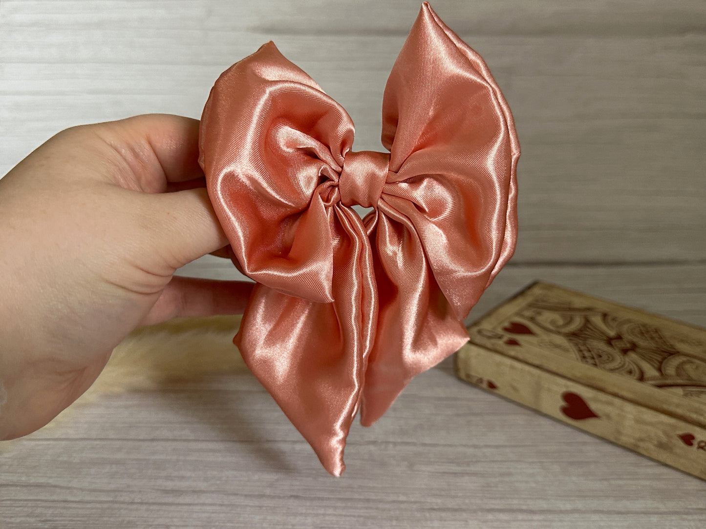 A hand holds the Crafts by Kate Silk Sailor Bow Clip in blush pink, its long satin tails elegantly draped against the wooden background. Next to it, a charming wooden box decorated with a heart pattern adds a delightful touch to the scene.