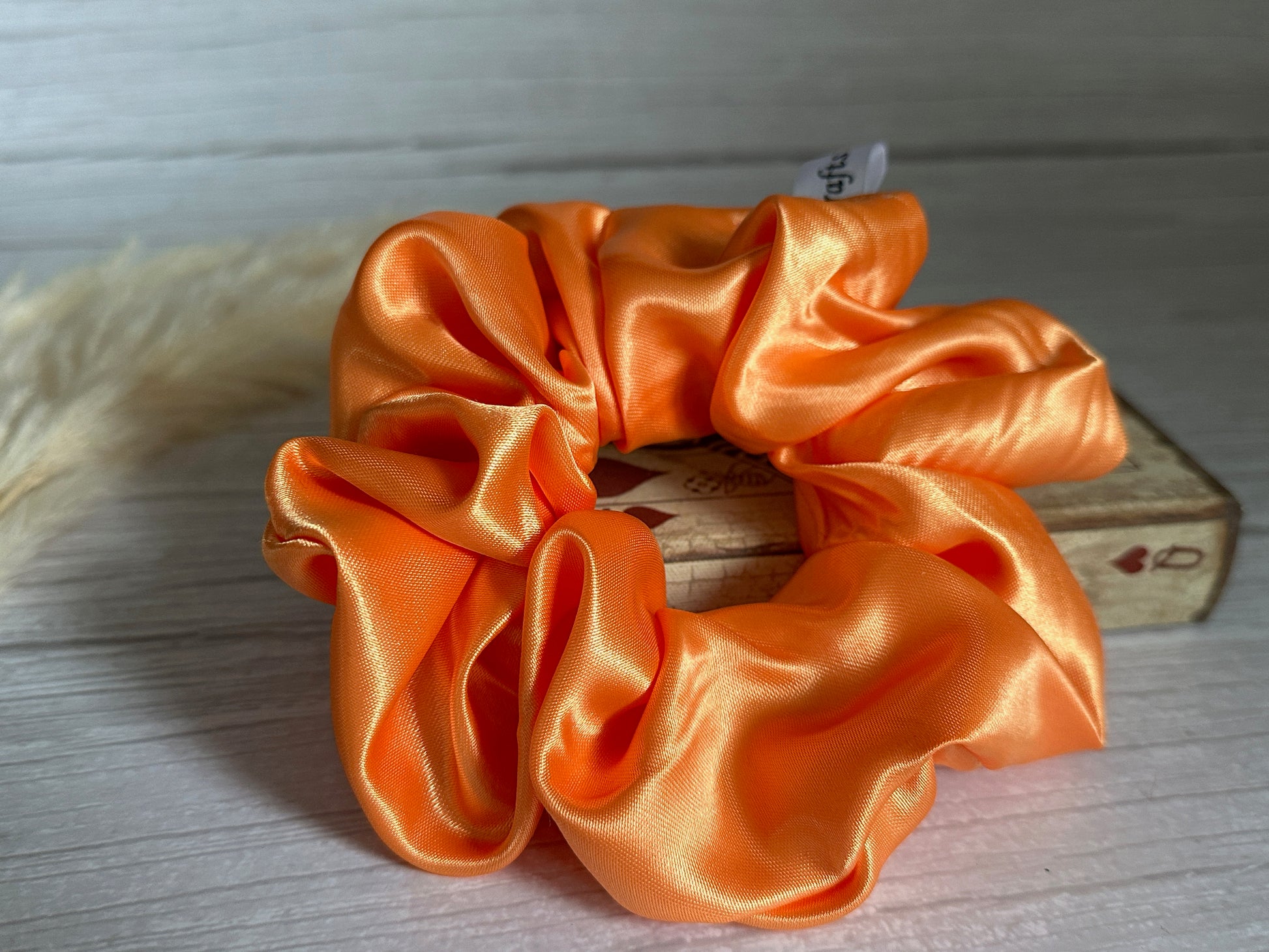 A Silk Extra Large Scrunchie - Bright Orange from Crafts by Kate is elegantly placed on a small wooden box, set against a light wooden surface. The glossy fabric, with its soft folds, creates a vibrant, textured appearance that exudes luxury.