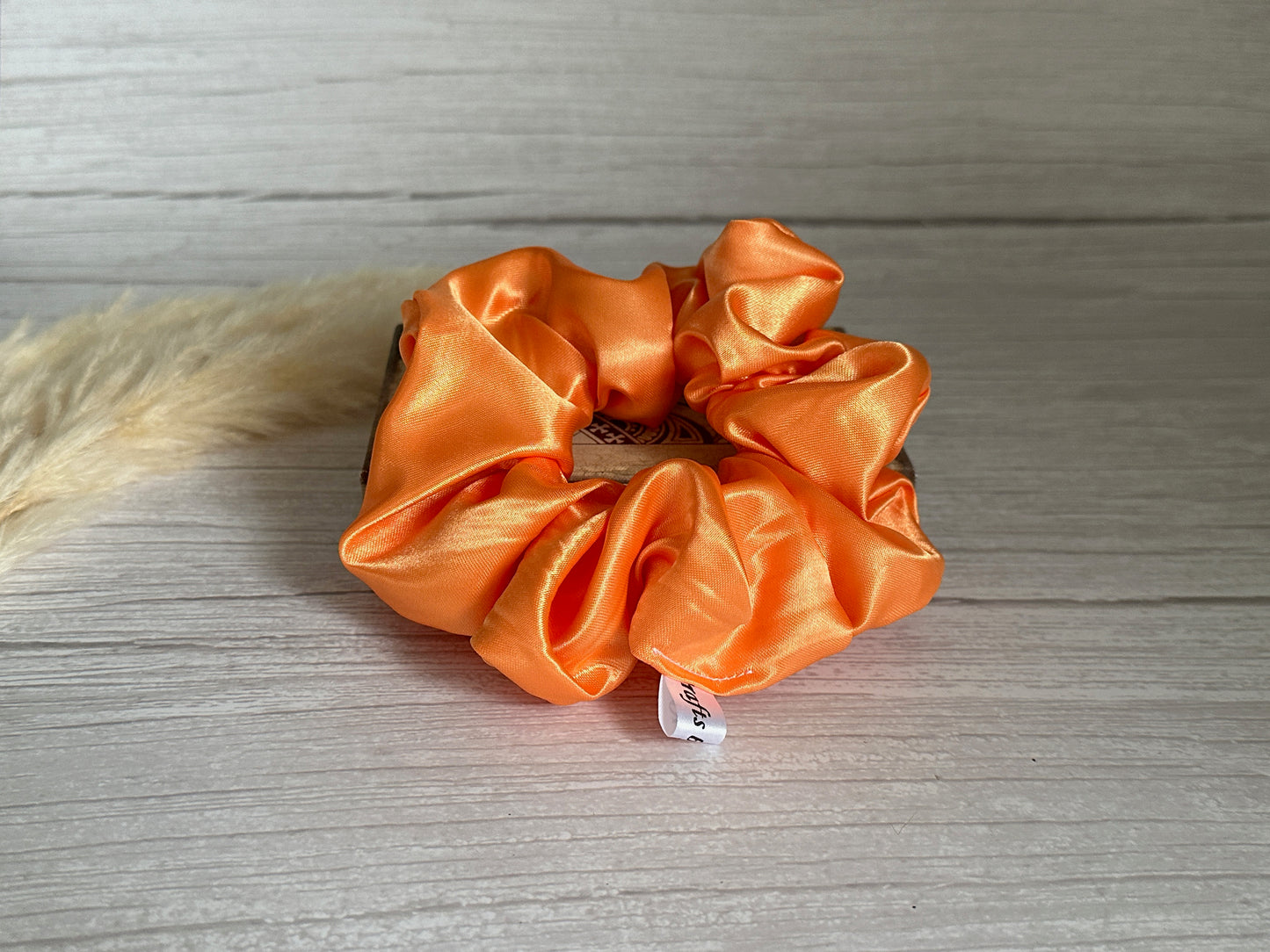 A Silk Extra Large Scrunchie in bright orange from Crafts by Kate, featuring a small white label, rests elegantly on a light wooden surface.