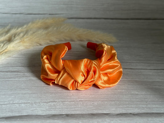 A Silk Knotted Headband in bright orange from Crafts by Kate, featuring a lustrous texture, is gracefully positioned on a wooden surface with delicate beige pampas grass adorning the background.