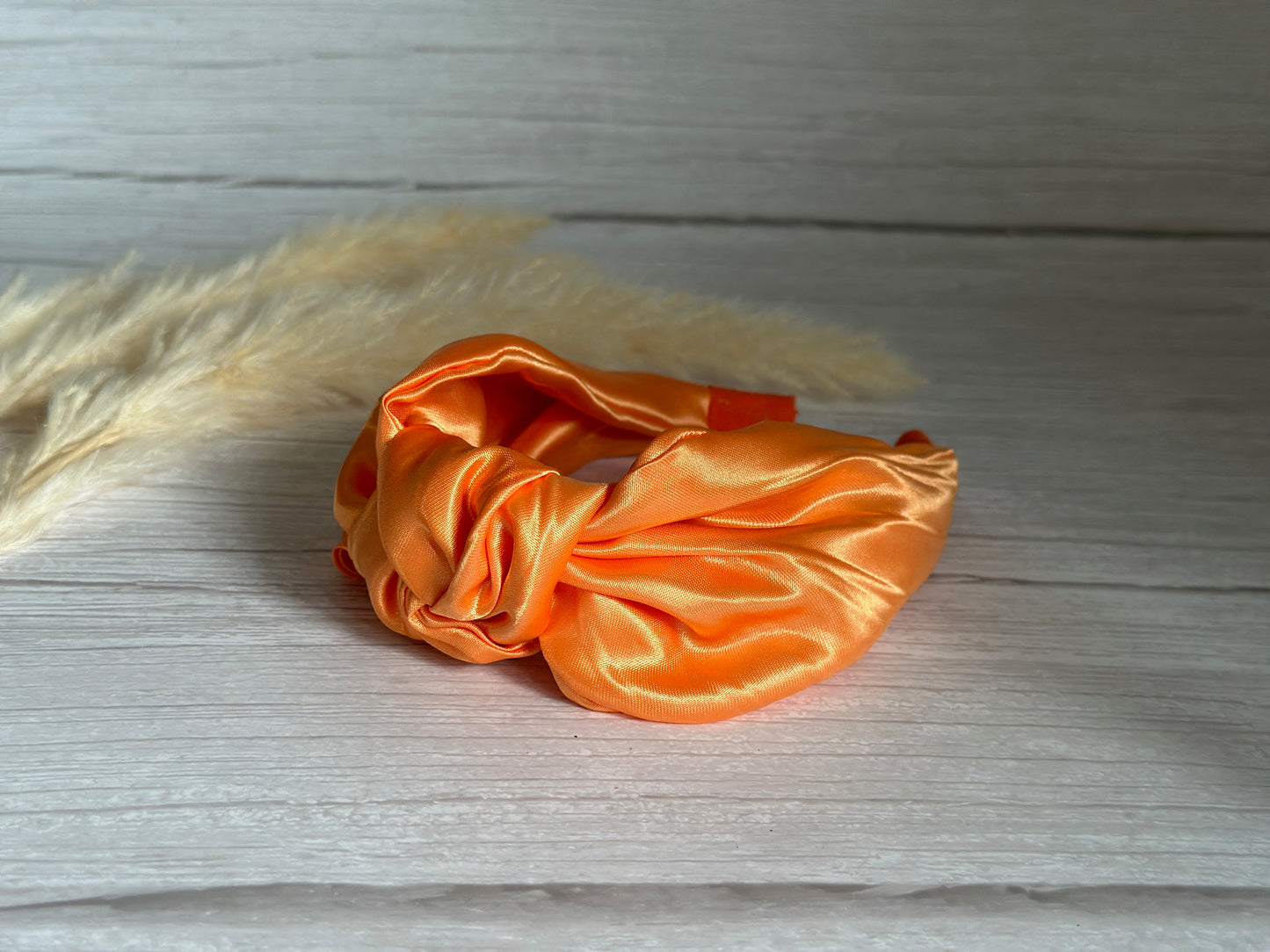 A vibrant Silk Knotted Headband in Bright Orange by Crafts by Kate lies on a light wooden surface, exuding handcrafted elegance as dried beige pampas grass gently peeks from the background.