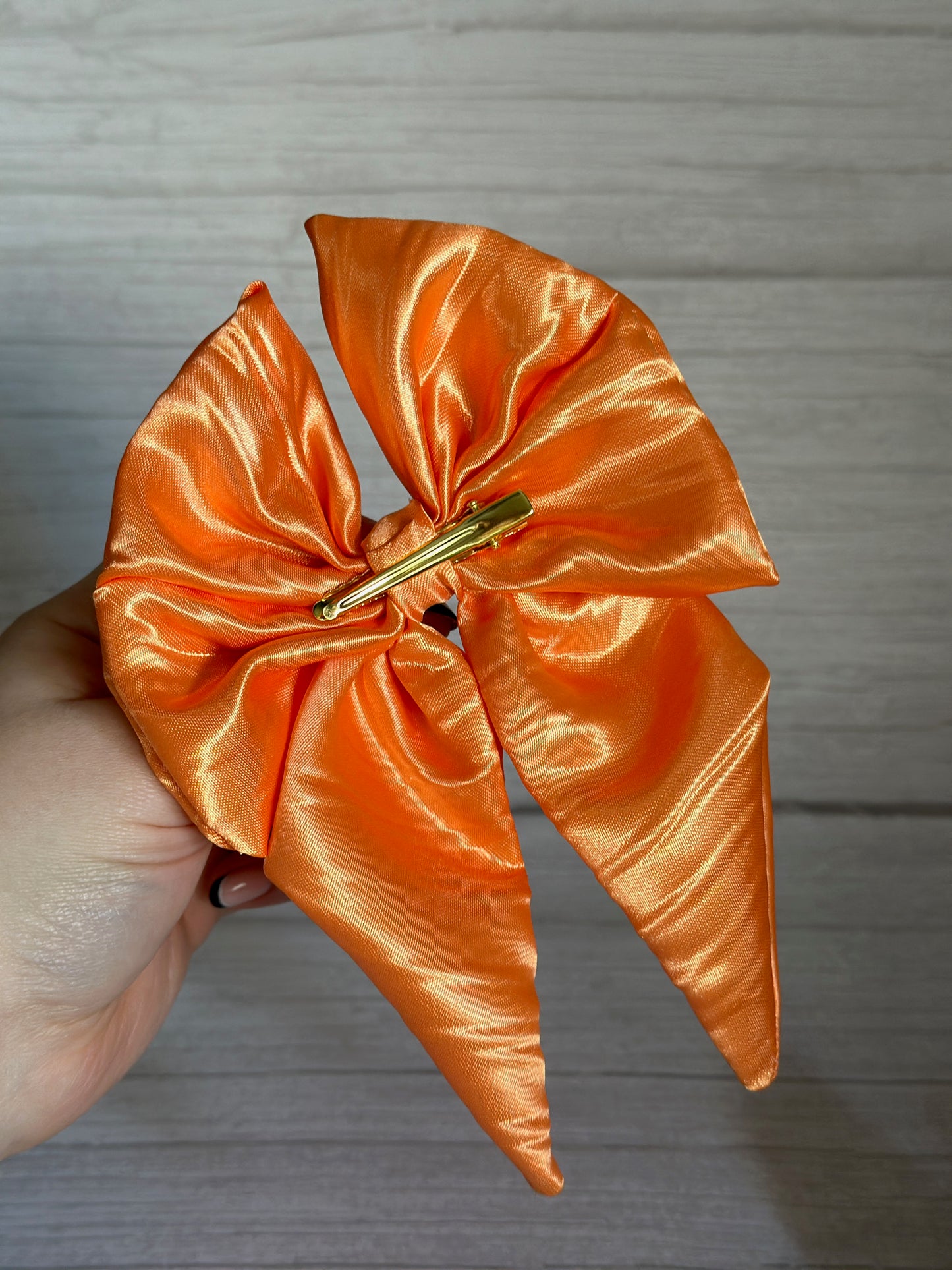 A hand delicately holds the Crafts by Kate Silk Sailor Bow Clip in bright orange, featuring a shiny satin finish and a metal clip, against a wooden background, exuding luxury.