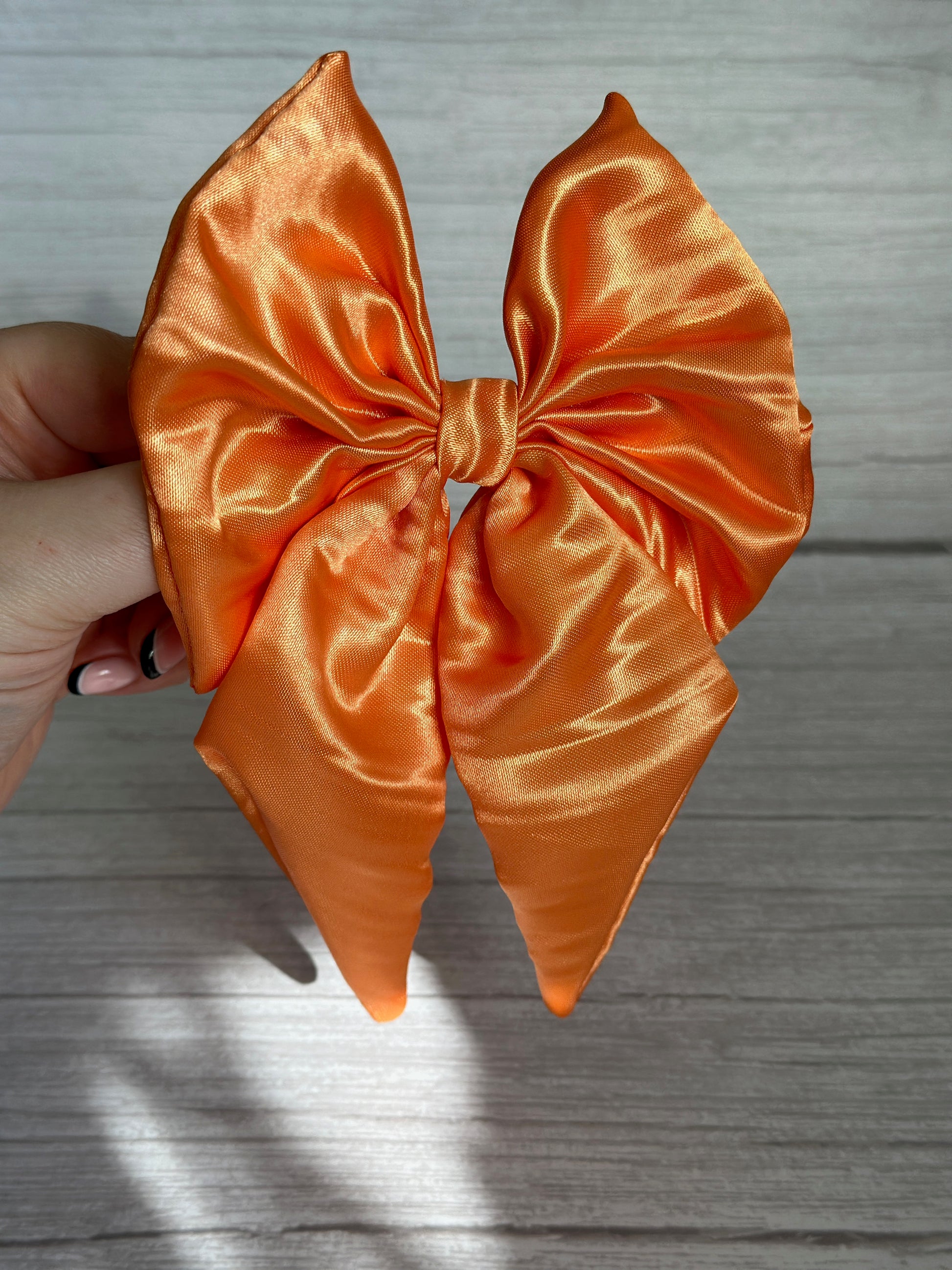 A hand holds the exquisite Silk Sailor Bow Clip - Bright Orange by Crafts by Kate against a light gray wood background. The large bow, made of lustrous silk and featuring long, pointed tails, radiates elegance.