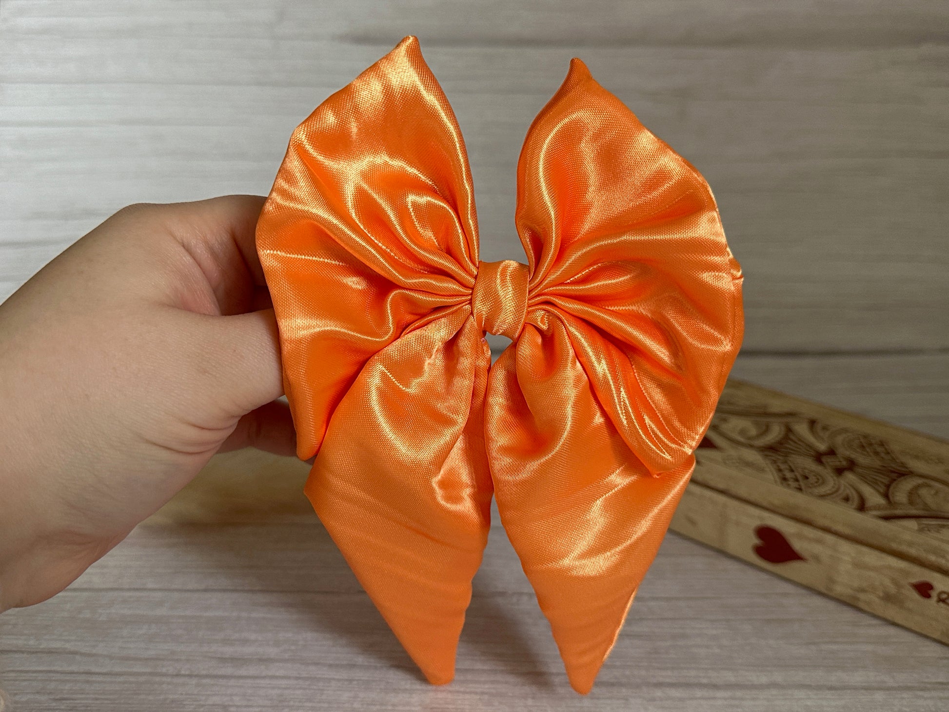 A hand holds the Crafts by Kate Silk Sailor Bow Clip in a luxurious and bright orange with a satin finish that evokes handcrafted silk. The background showcases a light wood surface and a decorative wooden box adorned with heart designs.