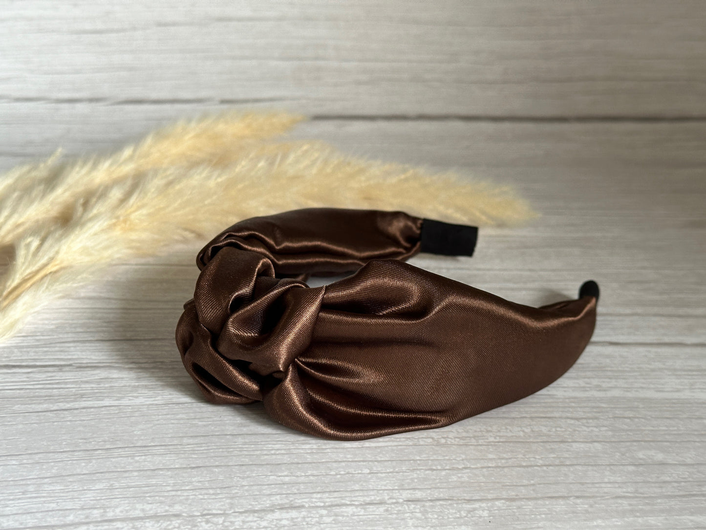 The Silk Knotted Headband in Chestnut Brown by Crafts by Kate exudes luxury as it lays gracefully on a light wood surface. The background's delicate pampas grass enhances its elegance, making this accessory a definitive luxury statement.