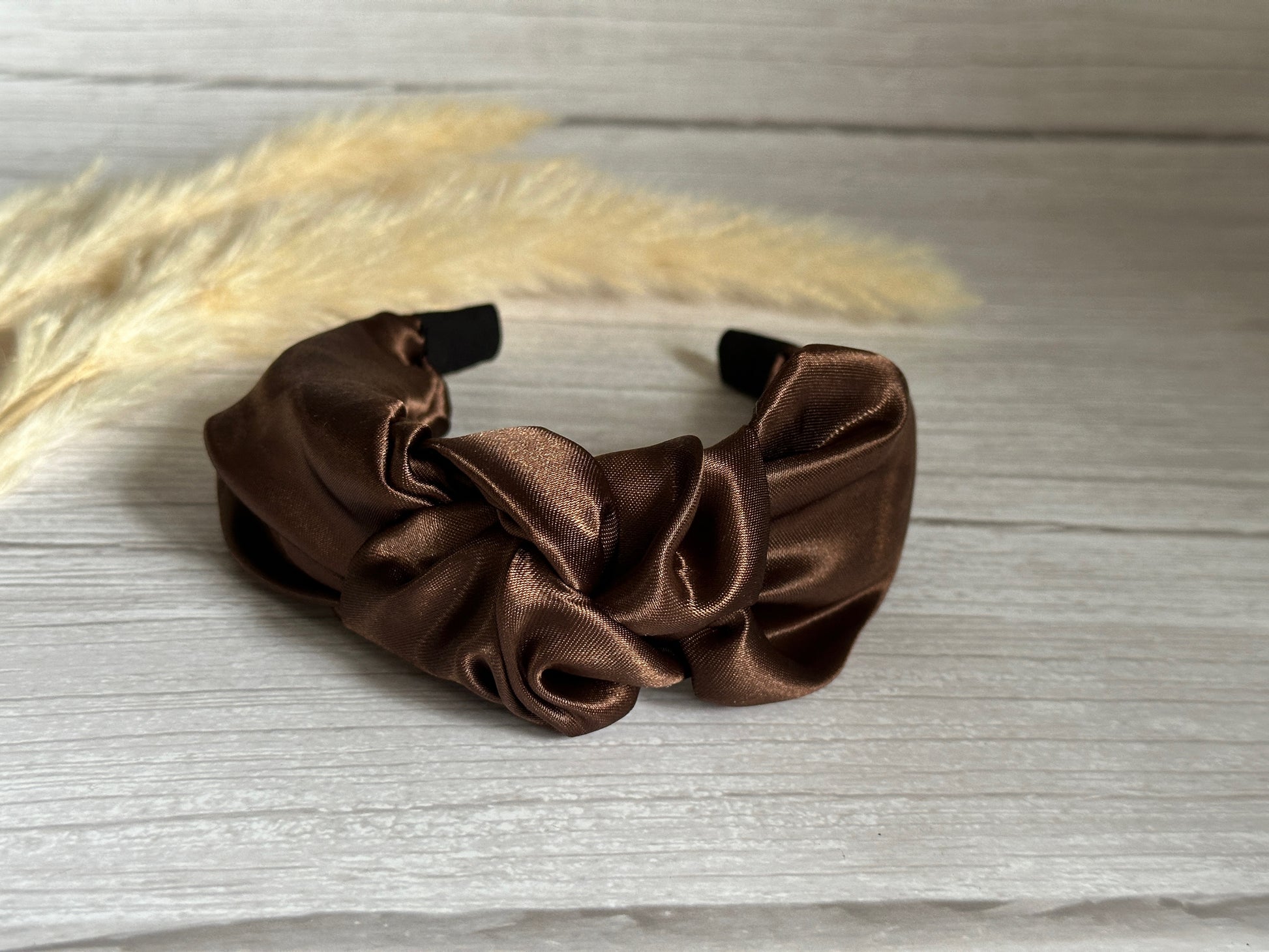 The Silk Knotted Headband in Chestnut Brown by Crafts by Kate serves as a luxurious statement piece, elegantly displayed on a light wooden surface. In the background, soft, beige dried grasses introduce a hint of nature's charm.