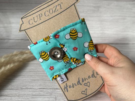 A hand holding a cardboard cutout labeled "Cotton Reusable Cup Cozy - Bumble Bee" by Crafts by Kate, showcasing a teal fabric cup sleeve adorned with bees, flowers, and a button. The handcrafted sleeve is labeled "Handmade" with hearts and features a small fabric tag on the side. This reusable accessory rests against a light wood surface in the background.